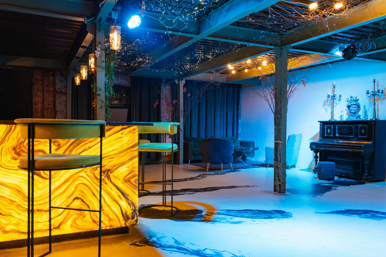 Арендовать Стилизованный двухэтажный лофт. Лофт-пространство с барной стойкой из светящегося камня, высокими стульями, черным пианино, креслами, светодиодным освещением на потолке, декор из веток, открытые балки, пол из светлого материала с темными полосами, приглушенное освещение, свечи.