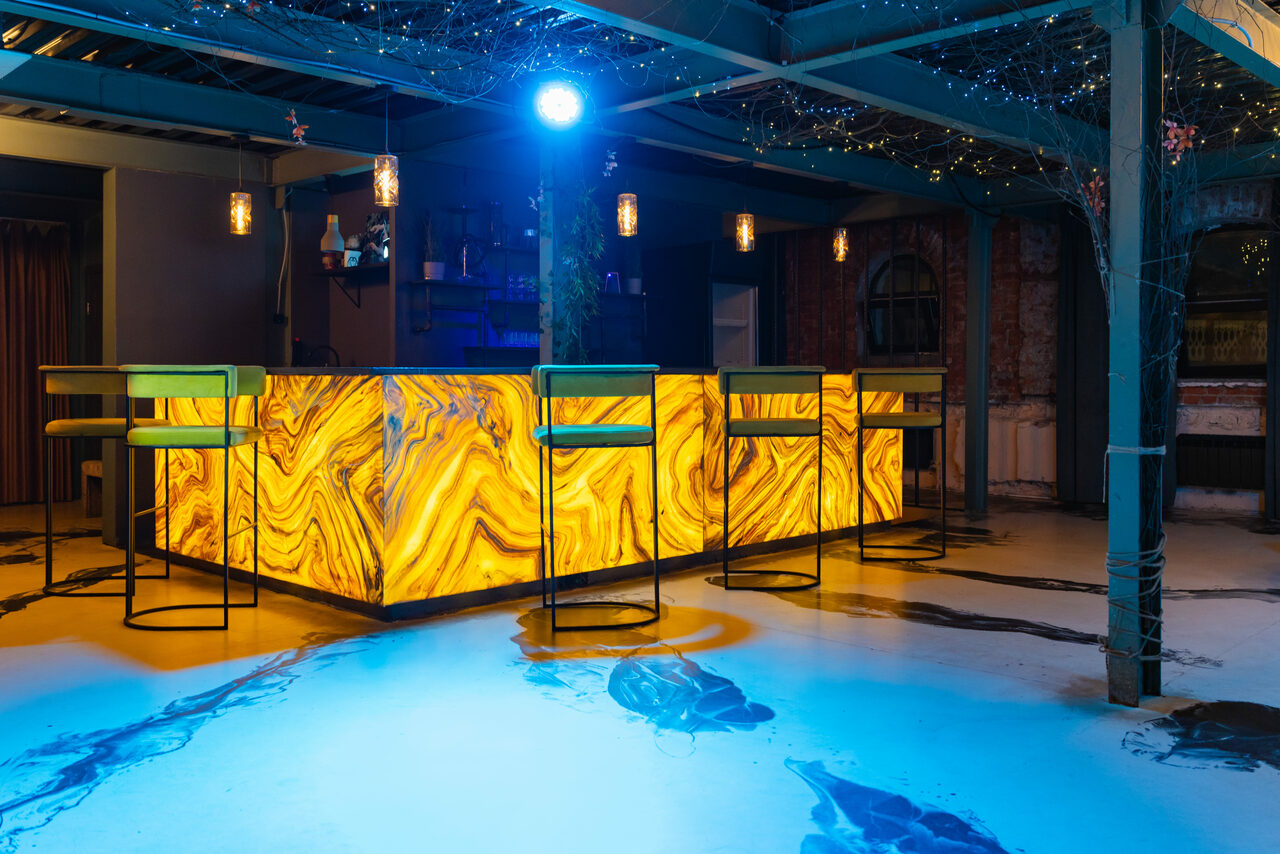 Арендовать Стилизованный двухэтажный лофт. Барная стойка с подсветкой янтарного цвета, высокие стулья, бетонный пол с синими прожекторами, подвесные светильники, открытые стеллажи с бутылками, декорированное помещение с металлическими конструкциями и светодиодными гирляндами.