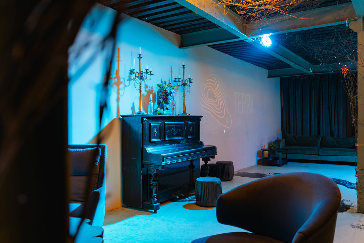 Арендовать Стилизованный двухэтажный лофт. Кабинет с фортепиано у стены, два кресла, пуфы, диван, подсвечники, декор на стене с абстрактным узором и надписью. Мягкое освещение с синим оттенком, бетонный пол, шторы на окне, видна часть лестницы и колонны.