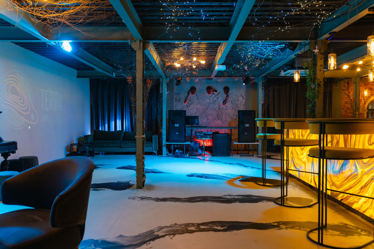 Арендовать Стилизованный двухэтажный лофт. Лофт-пространство с барной стойкой, высокими стульями, мягкими креслами, диваном, колонками и пианино. Потолок с декоративными элементами и гирляндами. Стены белые, пол светлый с темными узорами. Освещение теплое и холодное. На стене проекция.