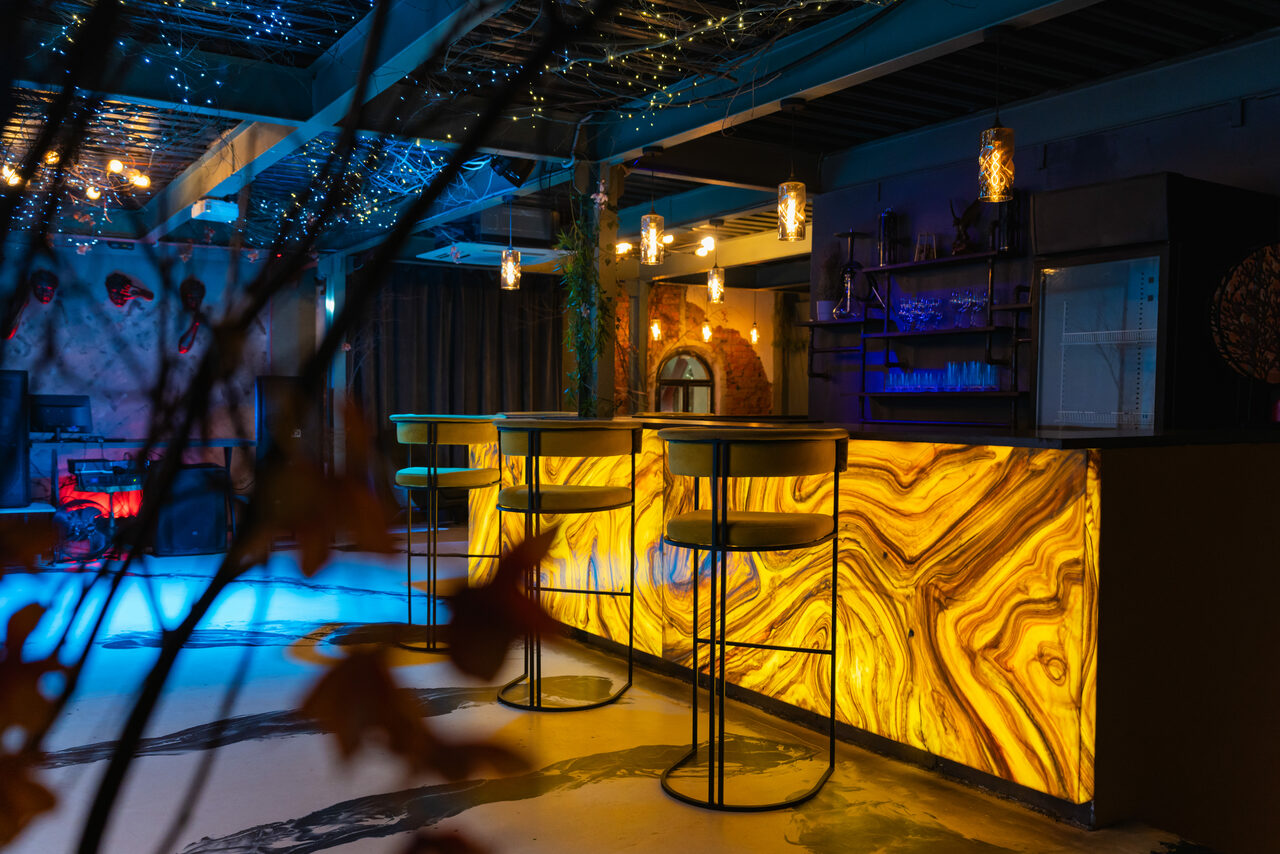 Арендовать Стилизованный двухэтажный лофт. Барное пространство с подсвеченной стойкой в мраморном стиле и высокими стульями. Принт из листьев на переднем плане. Разноцветная подсветка, висячие светильники. Пол — светлый, бетонный. Потолок с декоративными гирляндами. Стойка с полками и микроволновкой.