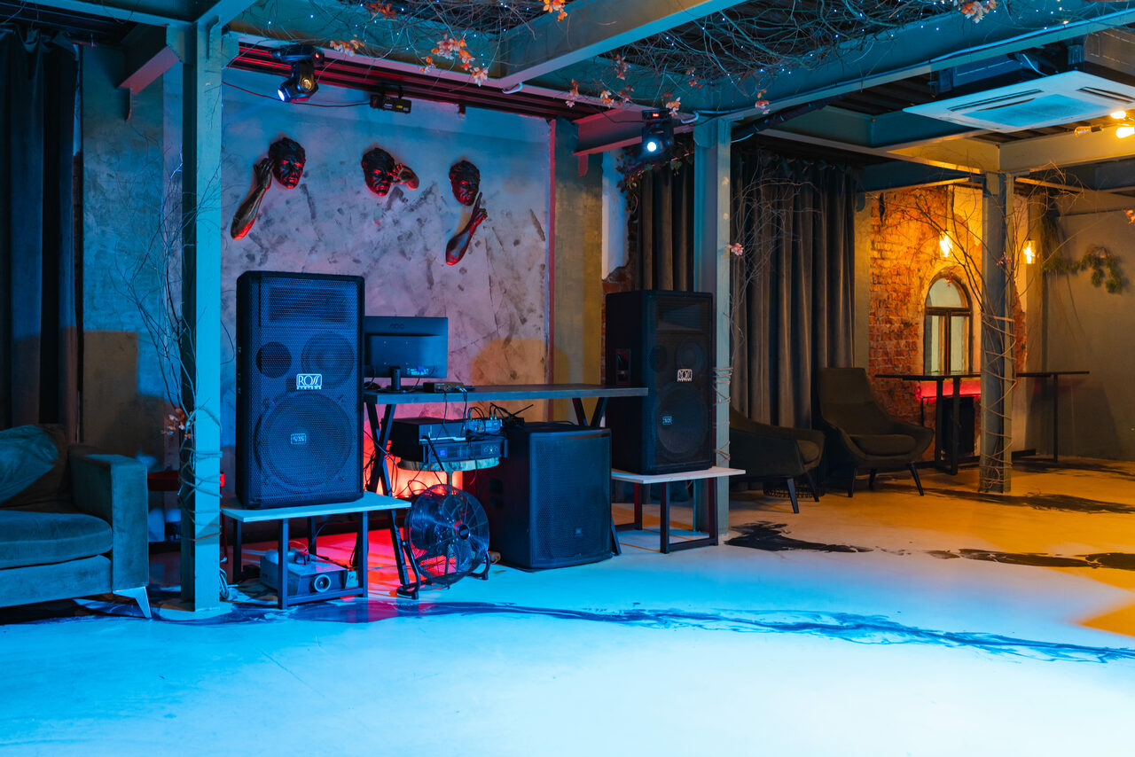 Арендовать Стилизованный двухэтажный лофт. Просторная комната с индустриальным дизайном, большая аудиосистема, диван, кресла, бетонный пол, кирпичные стены, декоративные маски на стене, яркое разноцветное освещение, металлические балки на потолке, кондиционер.