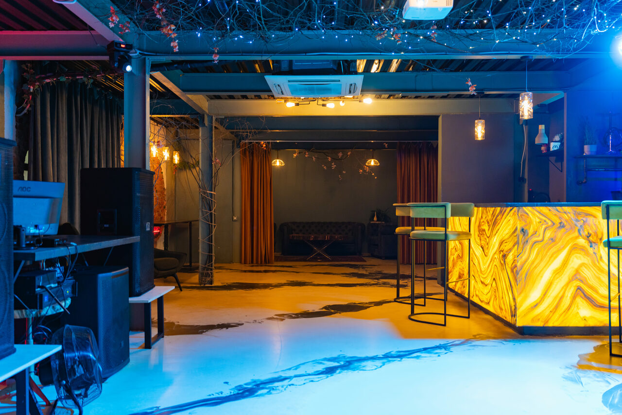Арендовать Стилизованный двухэтажный лофт. Пространство с барной стойкой, высокими стульями, аудиотехникой и диваном. Яркое декоративное освещение, натяжные потолки с декором. Пестрый пол, темные занавески на фоне. Стены серые с элементами декора.