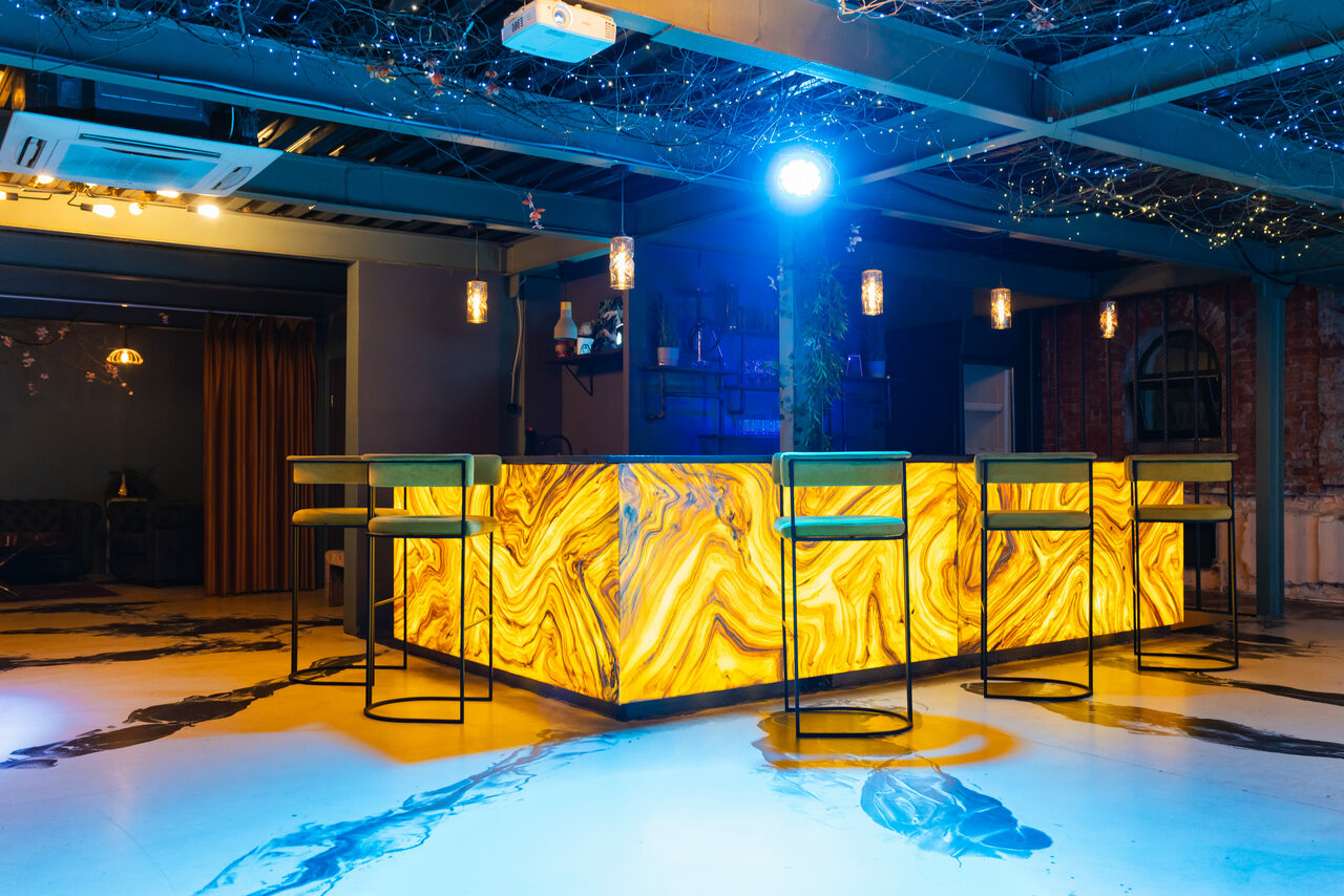 Арендовать Стилизованный двухэтажный лофт. Современный бар с подсвеченной стойкой из желтого материала с текстурой камня. Высокие металлические барные стулья с обивкой. Потолок открытой конструкции, подвесные светильники. Пол светлый с темными узорами. Стены темные, кирпичная кладка.