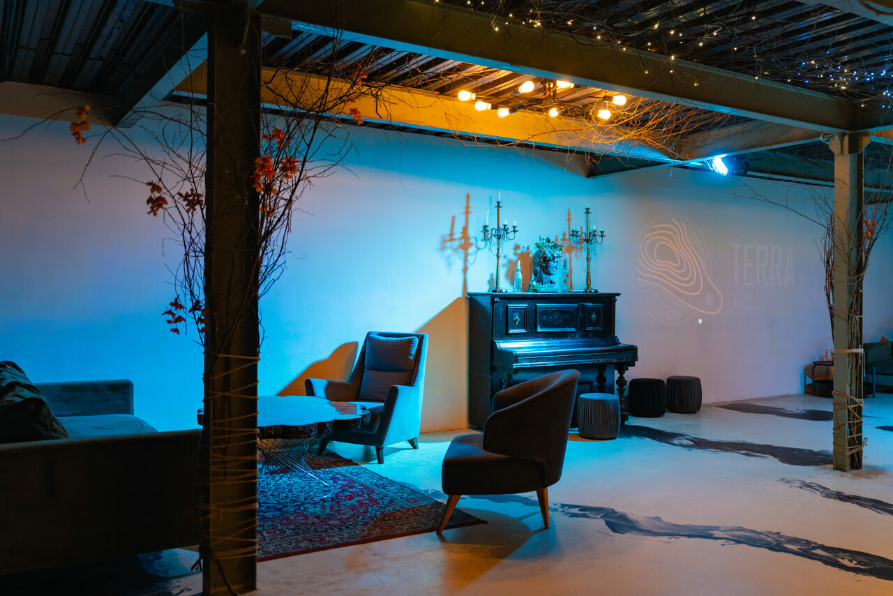 Арендовать Стилизованный двухэтажный лофт. Лофт-пространство с белыми стенами и бетонным полом, декорированное ветками. В комнате находится диван, несколько кресел, черное пианино и стол. Освещение теплое, потолок украшен светодиодными гирляндами. На стене надпись "TERR".