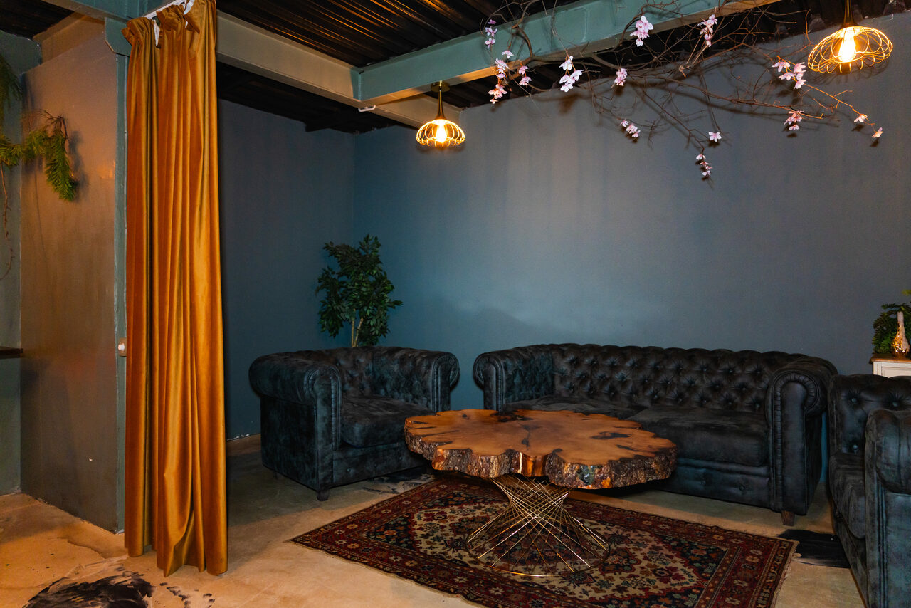 Арендовать Стилизованный двухэтажный лофт. Лофт-пространство с темной кожаной мягкой мебелью, большим деревянным столом с металлическими ножками, синими стенами и ковром с узором. Подвесные светильники теплого света и декоративные ветки с цветами. Занавески золотого цвета и зеленое растение в углу.