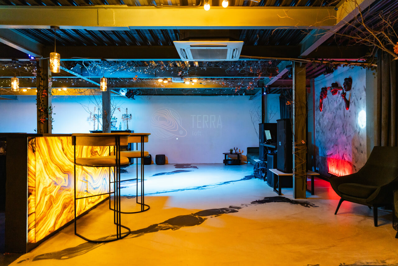 Арендовать Стилизованный двухэтажный лофт. Пространство в стиле лофт с барной стойкой, высокими стульями и диванами. Потолок с балками, подвесные светильники. Настенные декорации с растениями. Проекция на стену. Песочные и синие оттенки, пол с декоративными узорами.