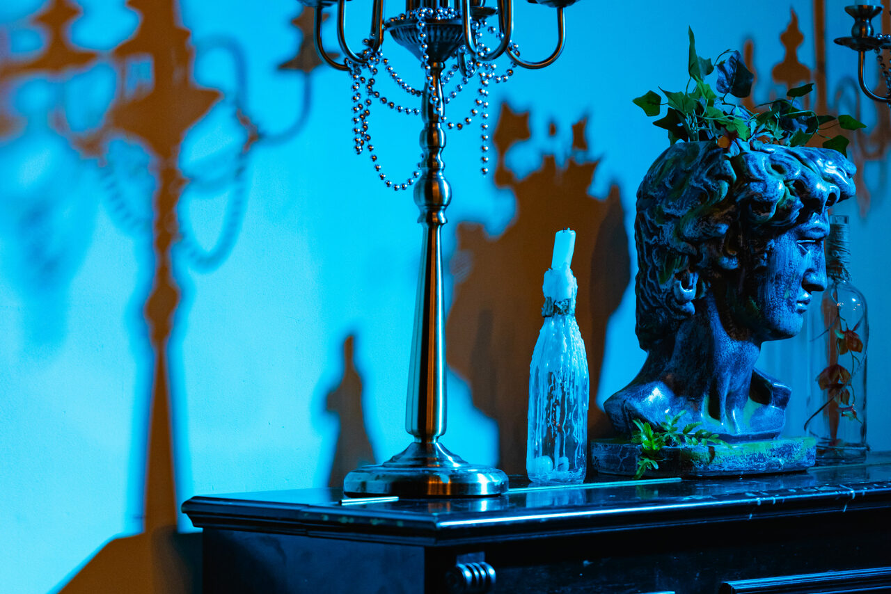 Арендовать Стилизованный двухэтажный лофт. Классическое пространство синим и оранжевым освещением; виден черный камин, на котором стоят статуя головы, подсвечник с цепями и бутылка; зелень украшает основу статуи.