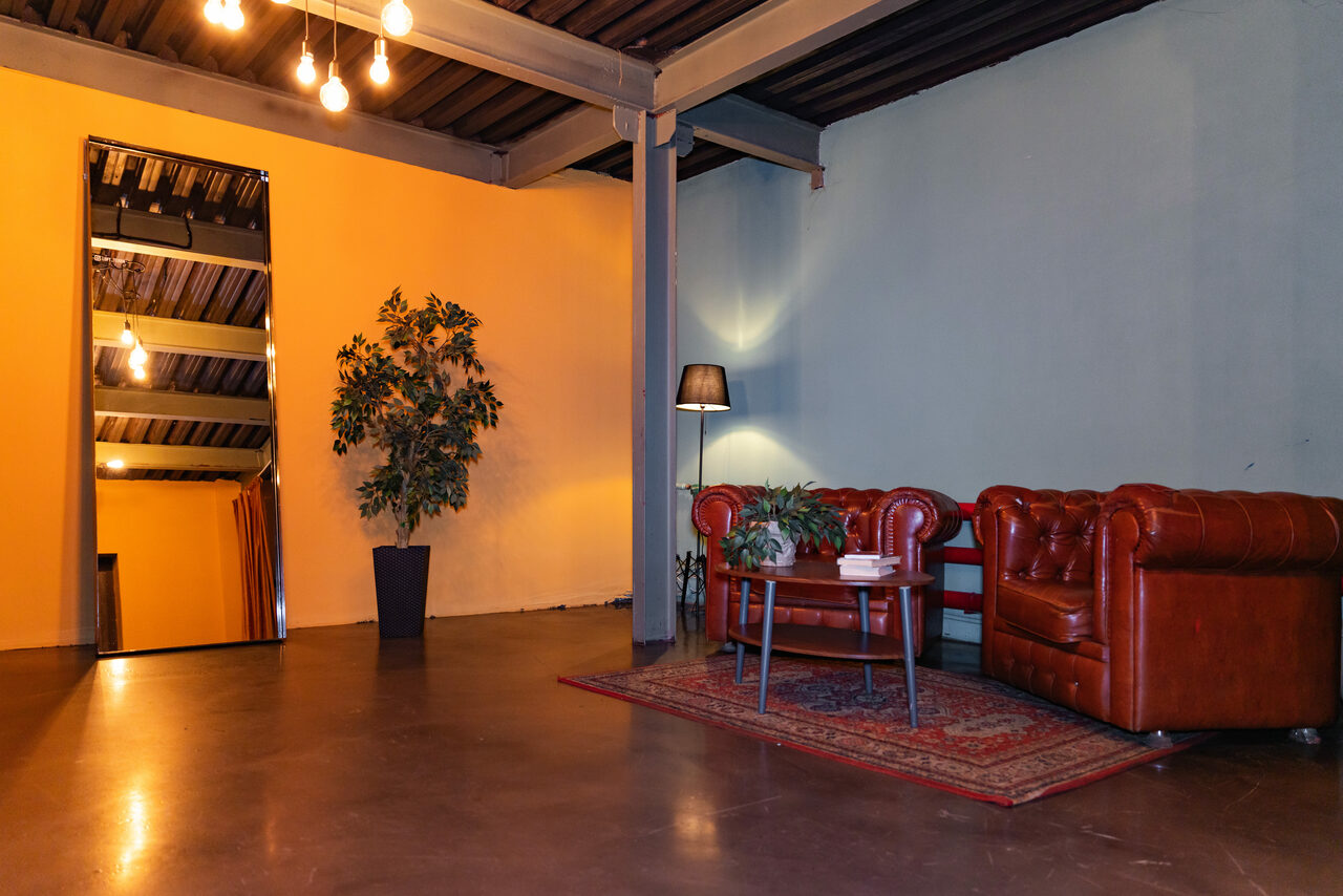 Арендовать Стилизованный двухэтажный лофт. Лофт-пространство с бетонным полом и высокими потолками. Красные кожаные диваны на ковре, рядом круглый стол. Настенное зеркало, декоративное растение, напольная лампа. Стены синие и оранжевые. Потолок с открытыми балками и подвесными лампами.