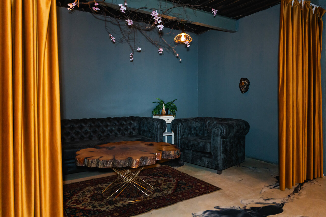 Арендовать Стилизованный двухэтажный лофт. Зона отдыха с двумя черными диванами, круглым столом с деревянной столешницей и металлическими ножками. Стены темно-синие, драпировка из золотистых штор. Потолок темный, центральное освещение и декоративные ветви с цветами. На полу ковер.