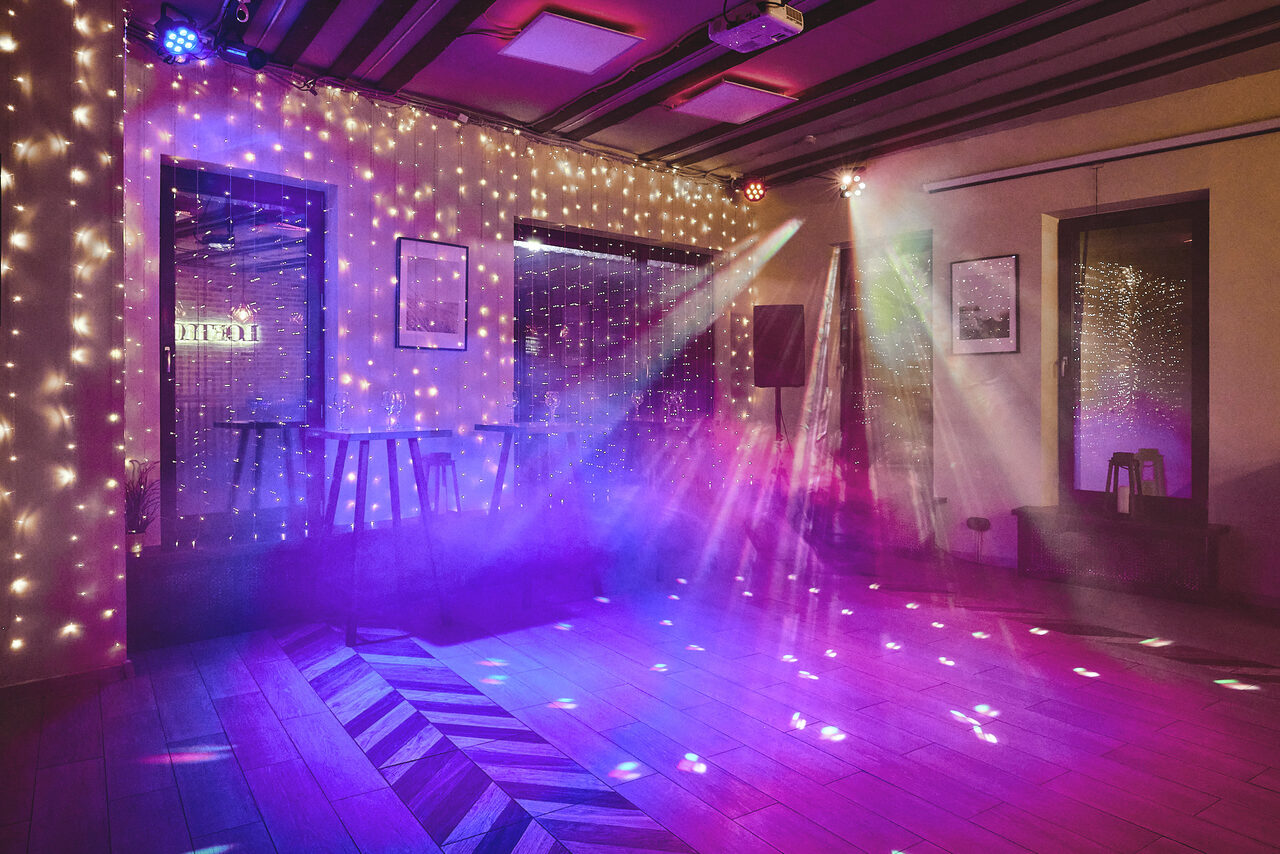 Арендовать Loft Lodge 6. Пространство с деревянным полом, светодиодными гирляндами на стенах, локальной подсветкой и световыми эффектами. Барные стулья у окна, стены бежевые, потолок с балками. Три светильника создают разноцветные лучи.