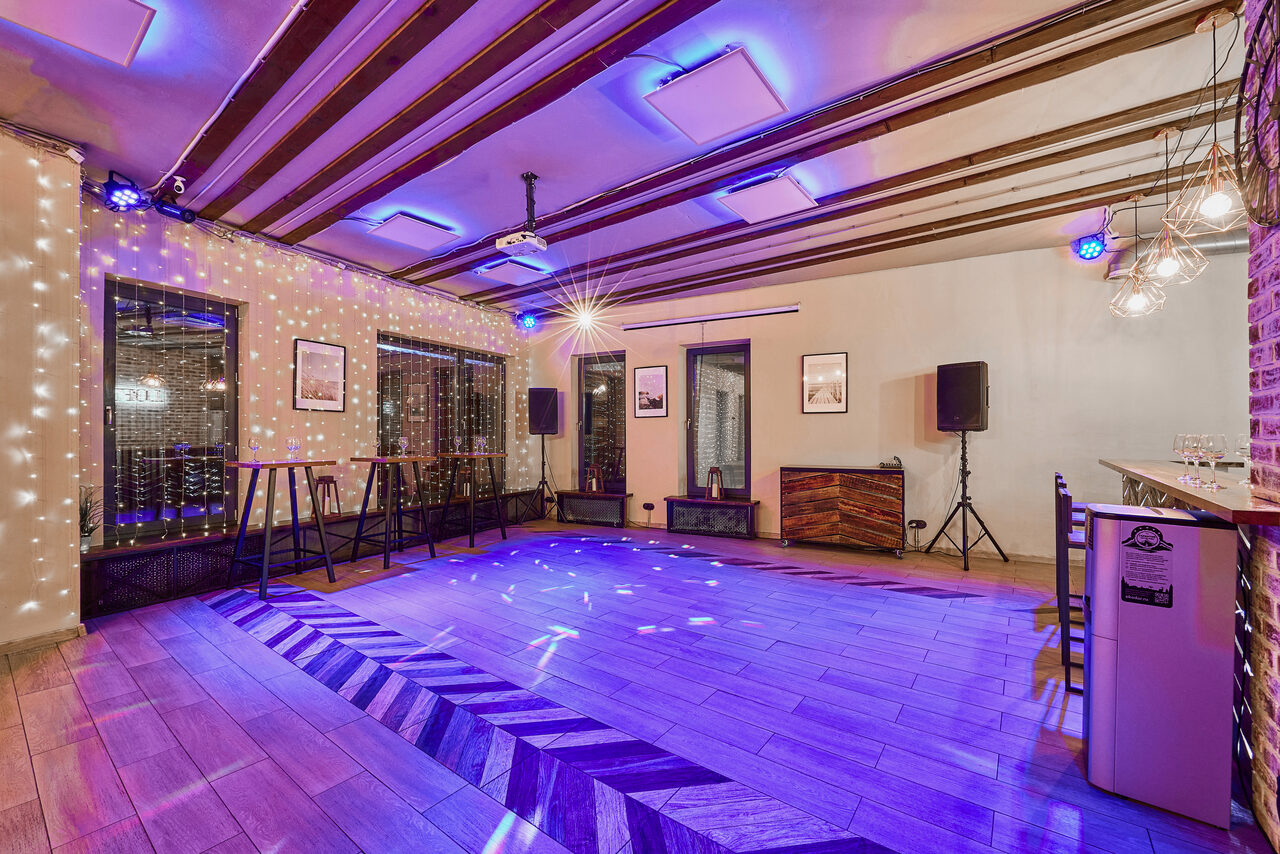 Арендовать Loft Lodge 6. Пространство с деревянным полом и потолочными балками, оборудовано колонками, барной стойкой и столами. Освещение фиолетовое и теплое, гирлянды и фотографии на стенах, на полу узор. кирпичная стена и декоративные светильники.