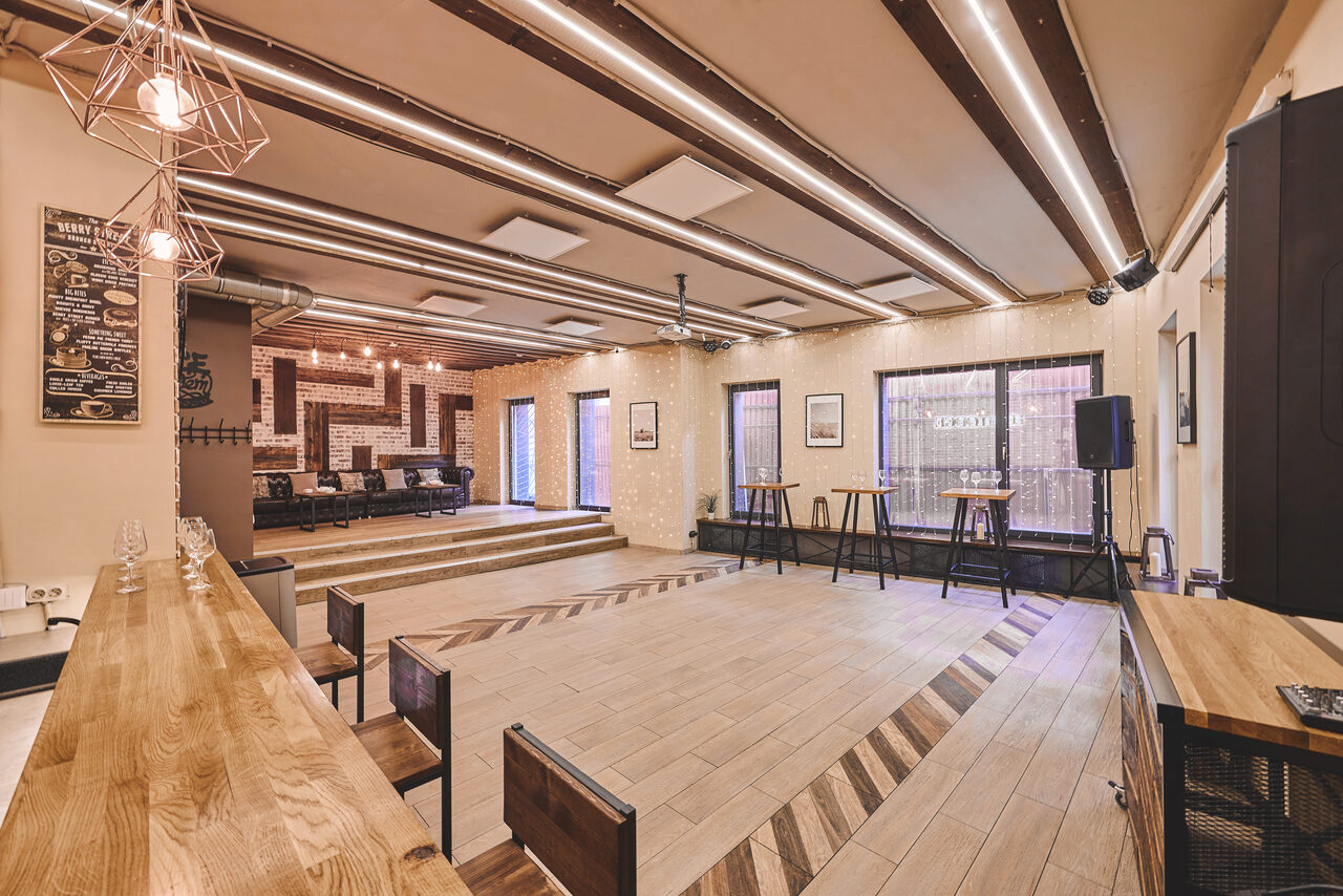 Арендовать Loft Lodge 6. Это просторное помещение с деревянным полом и ступенчатой планировкой. Имеется барная стойка с высокими стульями, сцена, столы и стулья. Потолок украшен балками и современными светильниками. Стены светлые, декорированы картинами. Надписи на меню слева от сцены.