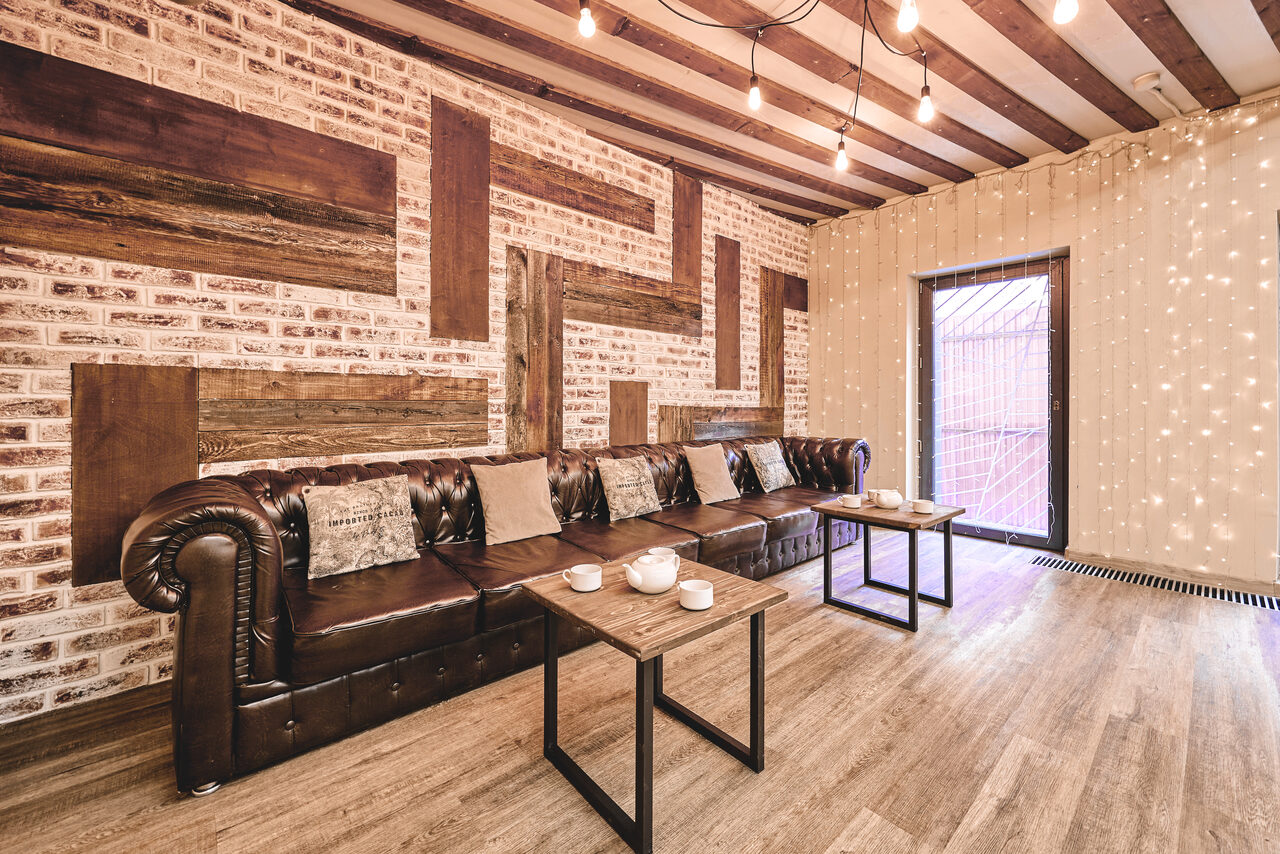 Арендовать Loft Lodge 6. Гостиная с кирпичными стенами и деревянными балками на потолке. Большой коричневый кожаный диван с подушками вдоль стены. Два деревянных стола перед диваном. Подвесное освещение с лампочками в индустриальном стиле. Пол тёмного дерева, входная дверь.