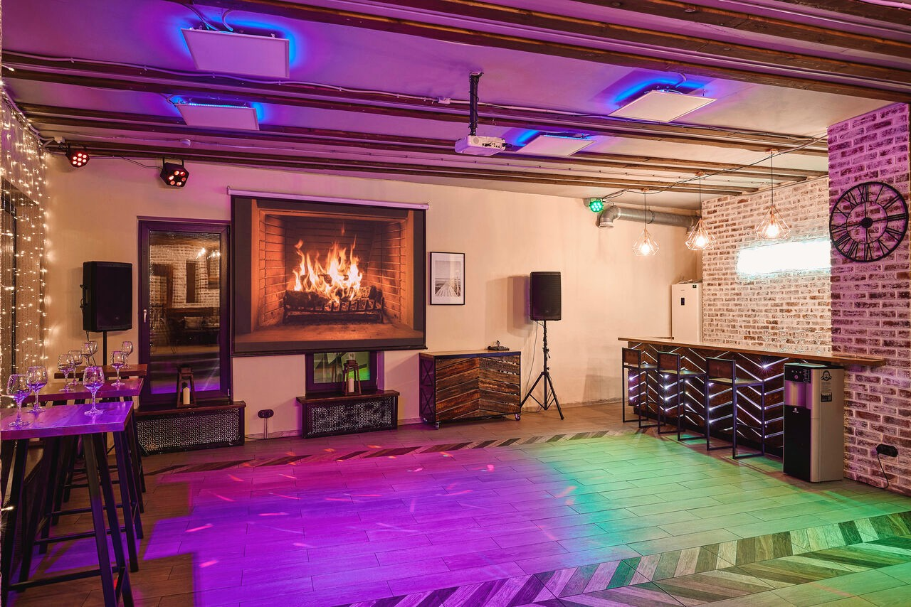 Арендовать Loft Lodge 6. Это внутреннее помещение, возможно, бар или лаунж-зона. Просторный пол выложен деревянными панелями. На стене изображение камина, рядом аппаратура, столы и барная стойка из дерева и металла. Освещение разноцветное, неоновые и гирляндные акценты, кирпичная стена.