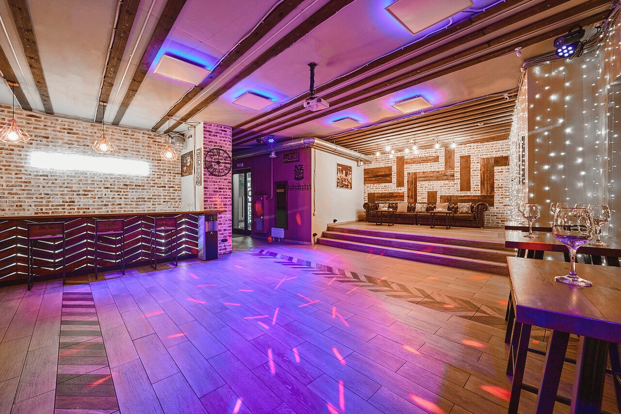 Арендовать Loft Lodge 6. Это просторное помещение с деревянным полом и кирпичными стенами. На потолке балки и декоративные панели с разноцветной подсветкой. Слева барная стойка, справа столы. В глубине сцена с диваном. Светодиодная гирлянда. Цвета теплые, сине-фиолетовое освещение.