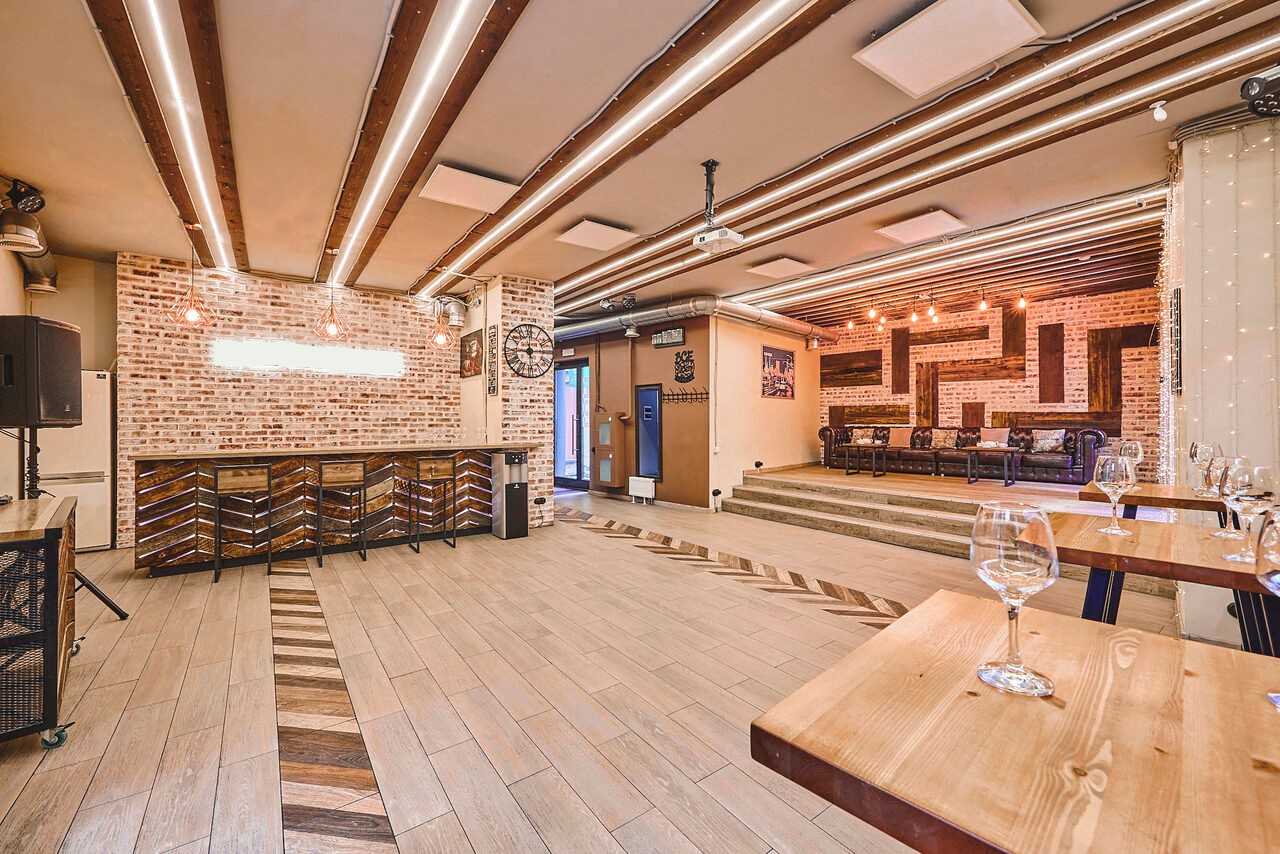 Арендовать Loft Lodge 6. На фотографии изображено просторное помещение с кирпичными стенами и деревянным полом. Видны деревянные столы и стулья, барная стойка с высокими стульями. Потолок украшен светильниками и деревянными балками. Чистое и аккуратное состояние.