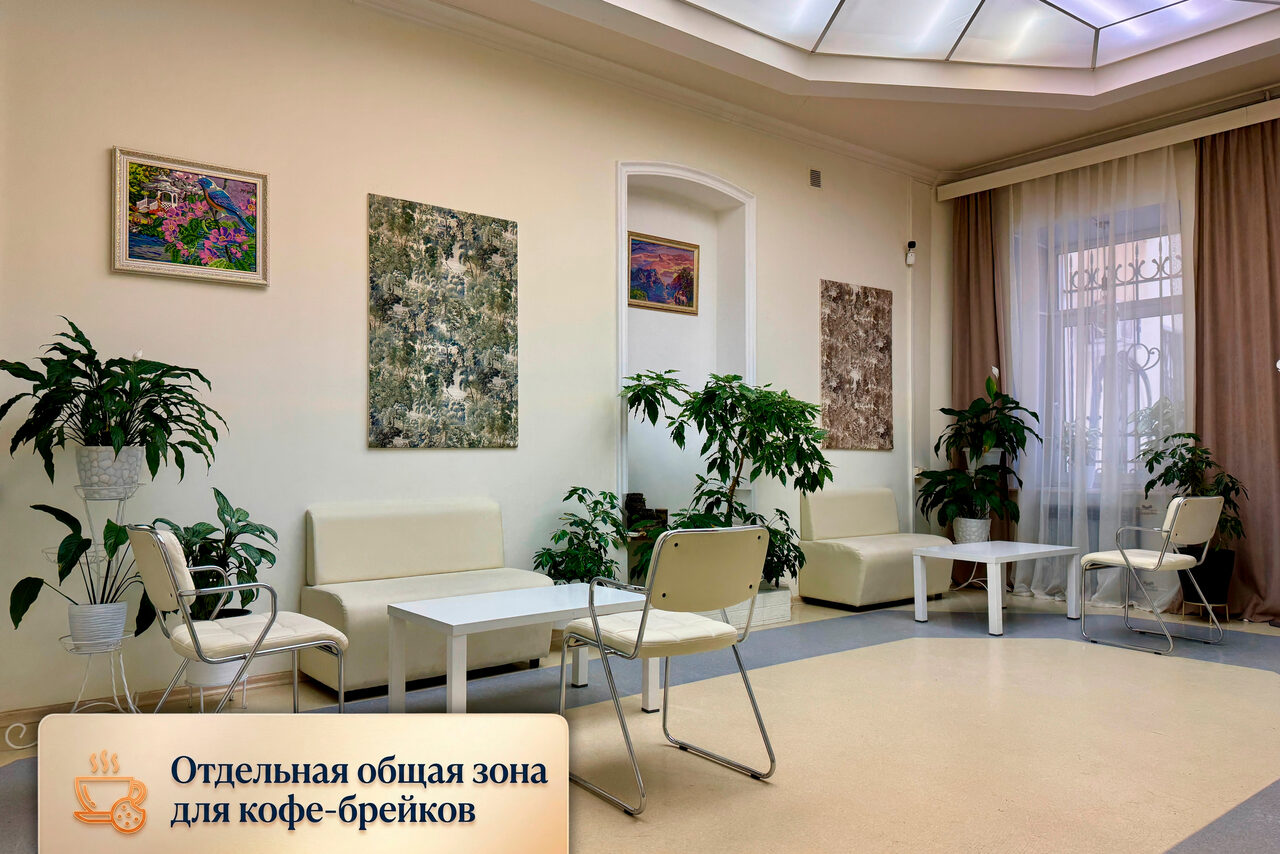 Арендовать Зал для мероприятий, тренингов, семинаров. Отдельная зона с мягкими бежевыми диванами и белыми столиками, окруженная зелеными растениями. Светлые стены украшены картинами. Естественное освещение через окно с белыми шторами. На стене текст: "Отдельная общая зона для кофе-брейков".
