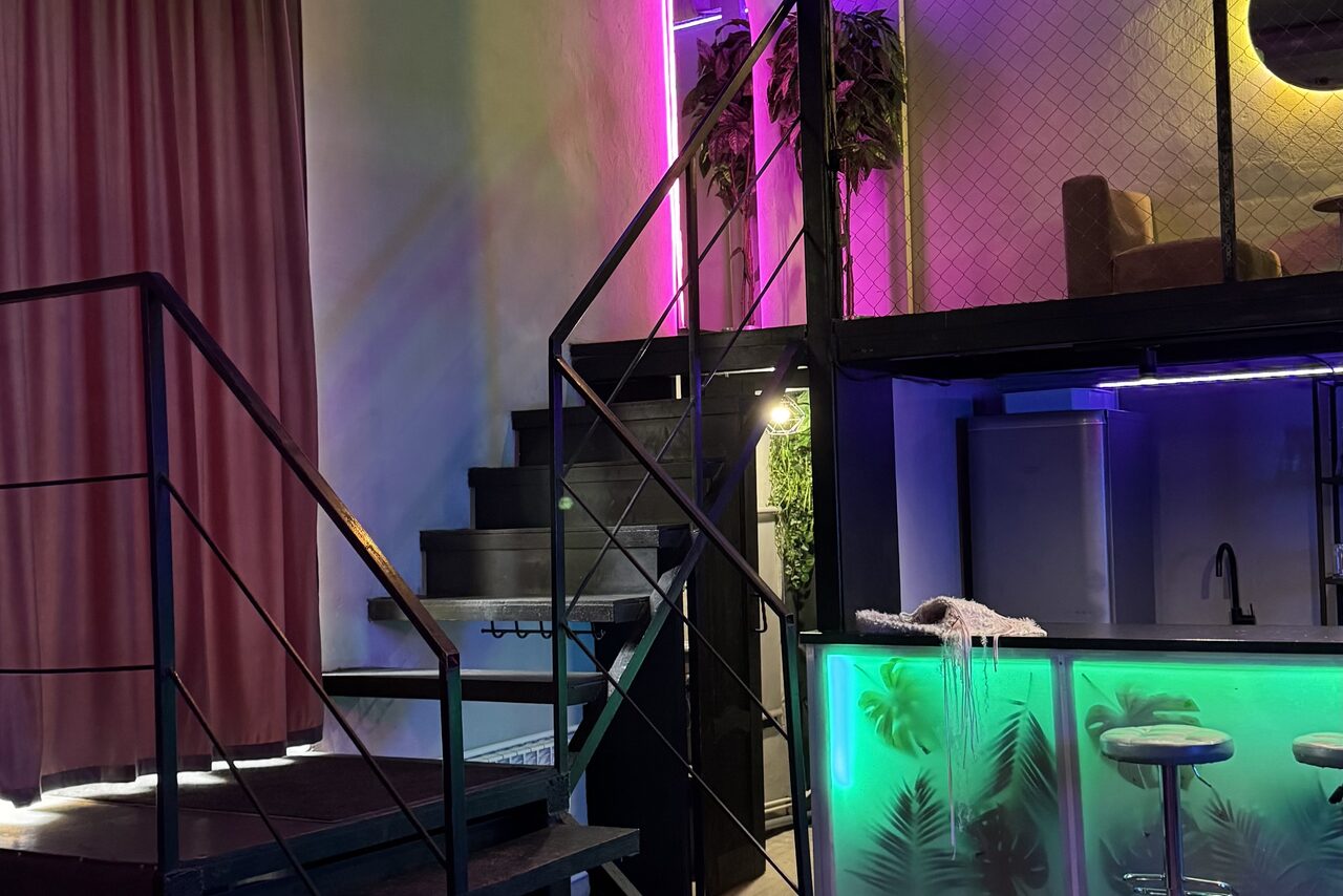 Арендовать The Portal (Портал). Тёмный неоновый лофт. Лофт-пространство с металлической лестницей, барной стойкой с неоновым освещением снизу и стульями. Стена украшена растениями. Верхний уровень с креслом и приглушенным светом. Цветовые акценты: розовый и зеленый. Штора и светодиодное освещение.