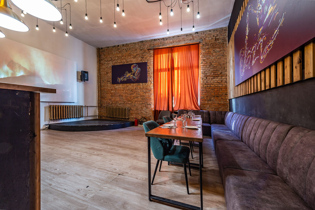 Арендовать Rubin Loft (Рубин Лофт). Зал Джаз. Просторное помещение с деревянными полами, кирпичной стеной и высоким потолком. Столы и стулья напольные, вдоль стены длинный диван. Оранжевые шторы на окне, освещение от подвесных ламп. На стенах декоративные панно. Маленькая сцена в углу.