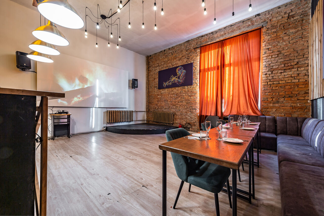 Арендовать Rubin Loft (Рубин Лофт). Зал Джаз. Многофункциональное помещение с кирпичными стенами и деревянным полом. Оранжевые шторы на окне, черные диваны. Столы с посудой, стулья. Потолочные светильники и проектор. Справа сцена, белая стена с экраном, мультимедийные колонки.