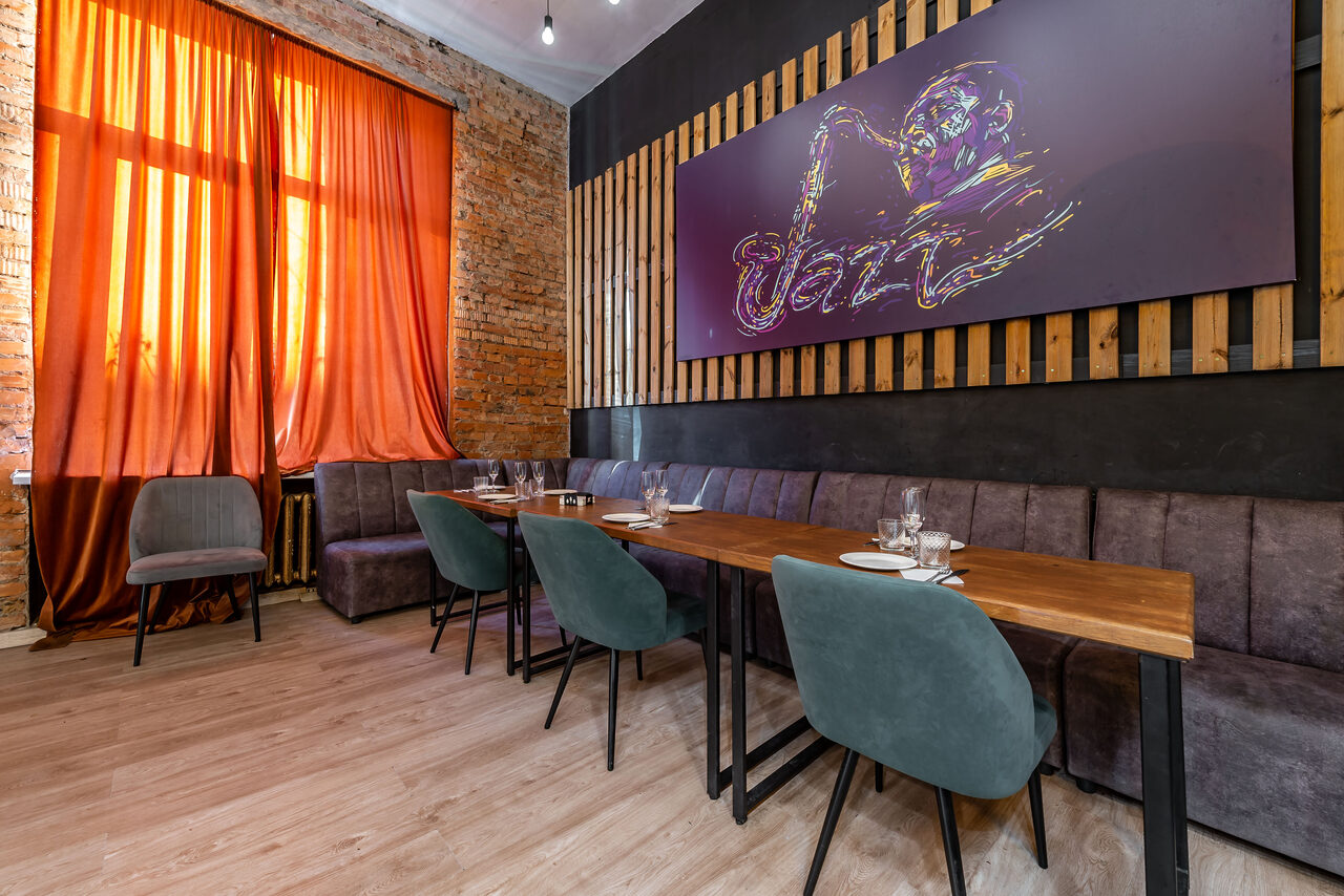Арендовать Rubin Loft (Рубин Лофт). Зал Джаз. Пространство с деревянным полом и кирпичными стенами. Деревянная мебель: длинные столы и мягкие стулья. На стене картина с надписью "Jazz". Красные шторы на окне справа. Естественное освещение через окно.