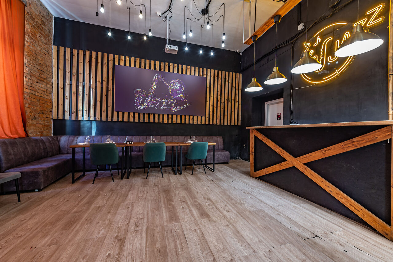 Арендовать Rubin Loft (Рубин Лофт). Зал Джаз. Просторное помещение с деревянным полом и кирпичной стеной, столы и стулья вдоль стены с деревянными панелями. Светильники на потолке. Неоновая надпись и изображение на стене. Барная стойка из темного дерева с крестом из светлого дерева.