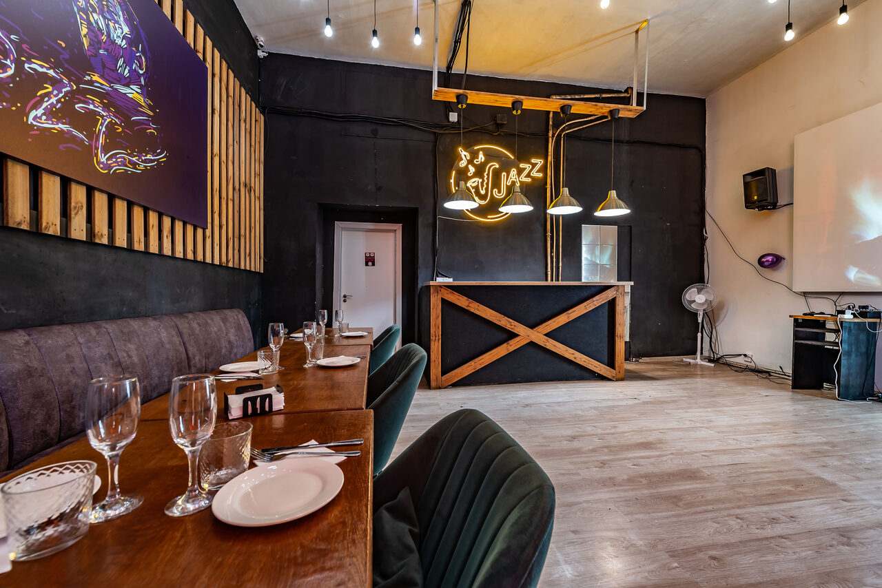 Арендовать Rubin Loft (Рубин Лофт). Зал Джаз. Просторное помещение с деревянными полами, столами и стульями. На стене неоновая вывеска с надписью "jazz". Освещение из подвесных светильников. Справа установлена аудиосистема. Потолок светлый, стена темная с декоративными элементами.