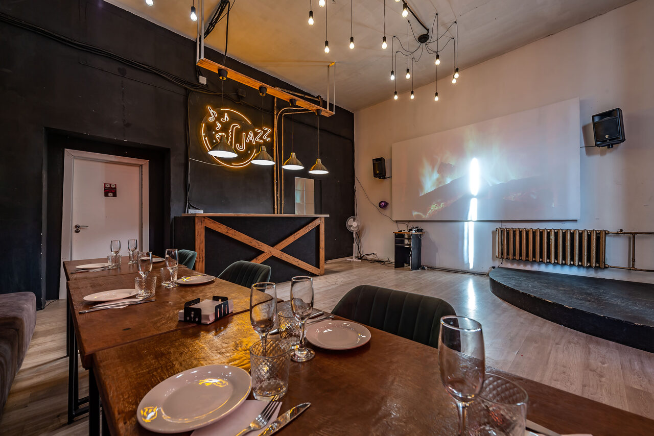Арендовать Rubin Loft (Рубин Лофт). Зал Джаз. Пространство кафе с деревянными столами, сервированными посудой и бокалами. На стене проекция изображения, рядом с ней аудиоколонка. Черные стены, декоративное освещение с лампами накаливания. Потолок светлый, пол деревянный. Барная стойка.