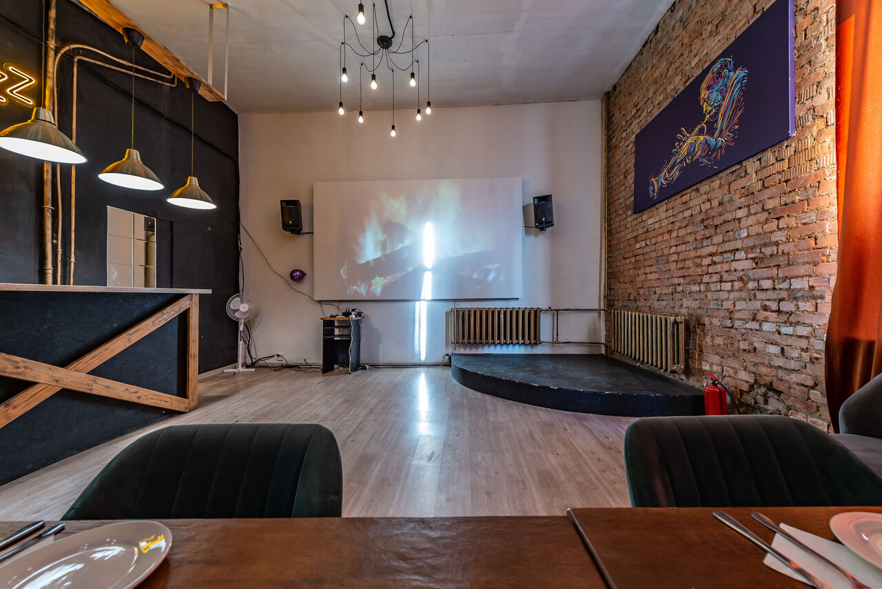 Арендовать Rubin Loft (Рубин Лофт). Зал Джаз. Пространство для мероприятий с барной стойкой и мягкими стульями. На стене кирпичная кладка и белая панель с проектором. Подвесные лампы над баром, люстра с лампами на потолке. Деревянные и металлические элементы, оранжевые шторы.