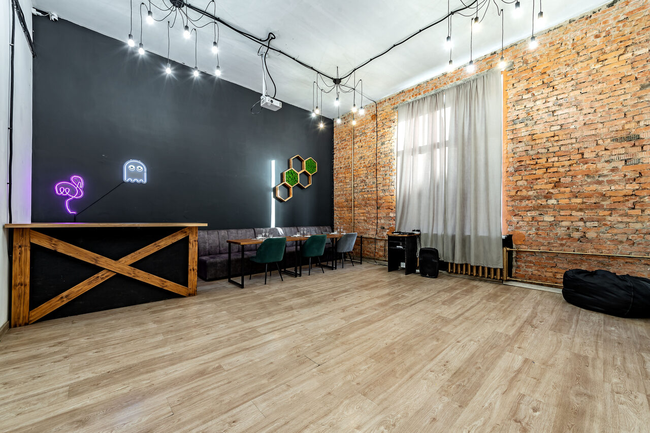 Арендовать Rubin Loft (Рубин Лофт). Лофт средний. Просторное помещение с кирпичными стенами и деревянным полом. Стена окрашена в чёрный с неоновым освещением. Есть барная стойка, столы и стулья, диван. Подвесное освещение. Большое окно с занавесками.