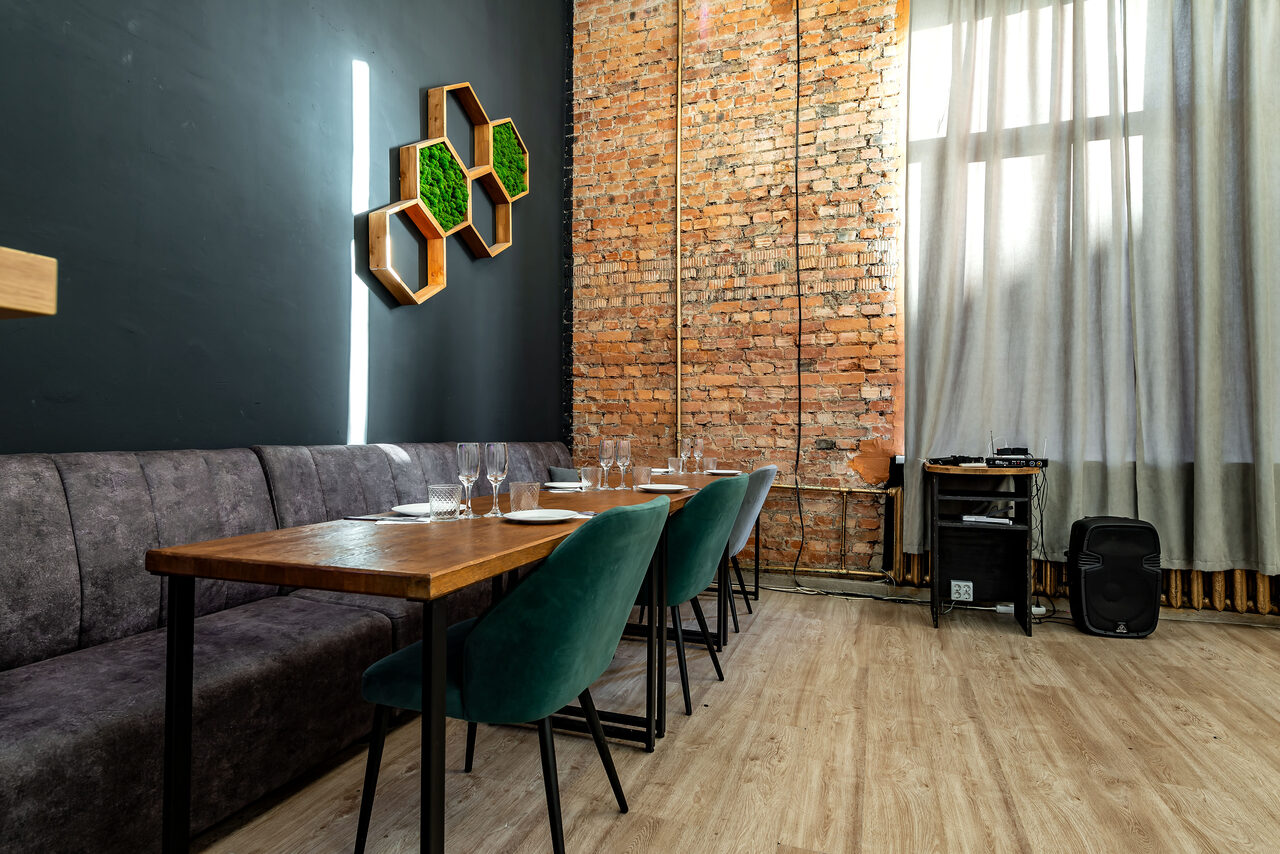 Арендовать Rubin Loft (Рубин Лофт). Лофт средний. Современное помещение с серой стеной и декоративными элементами из древесины в виде шестиугольников. Справа — кирпичная стена. У стены длинный стол, серая скамья и зеленые стулья. На полу светлое деревянное покрытие. Свет через большие окна.