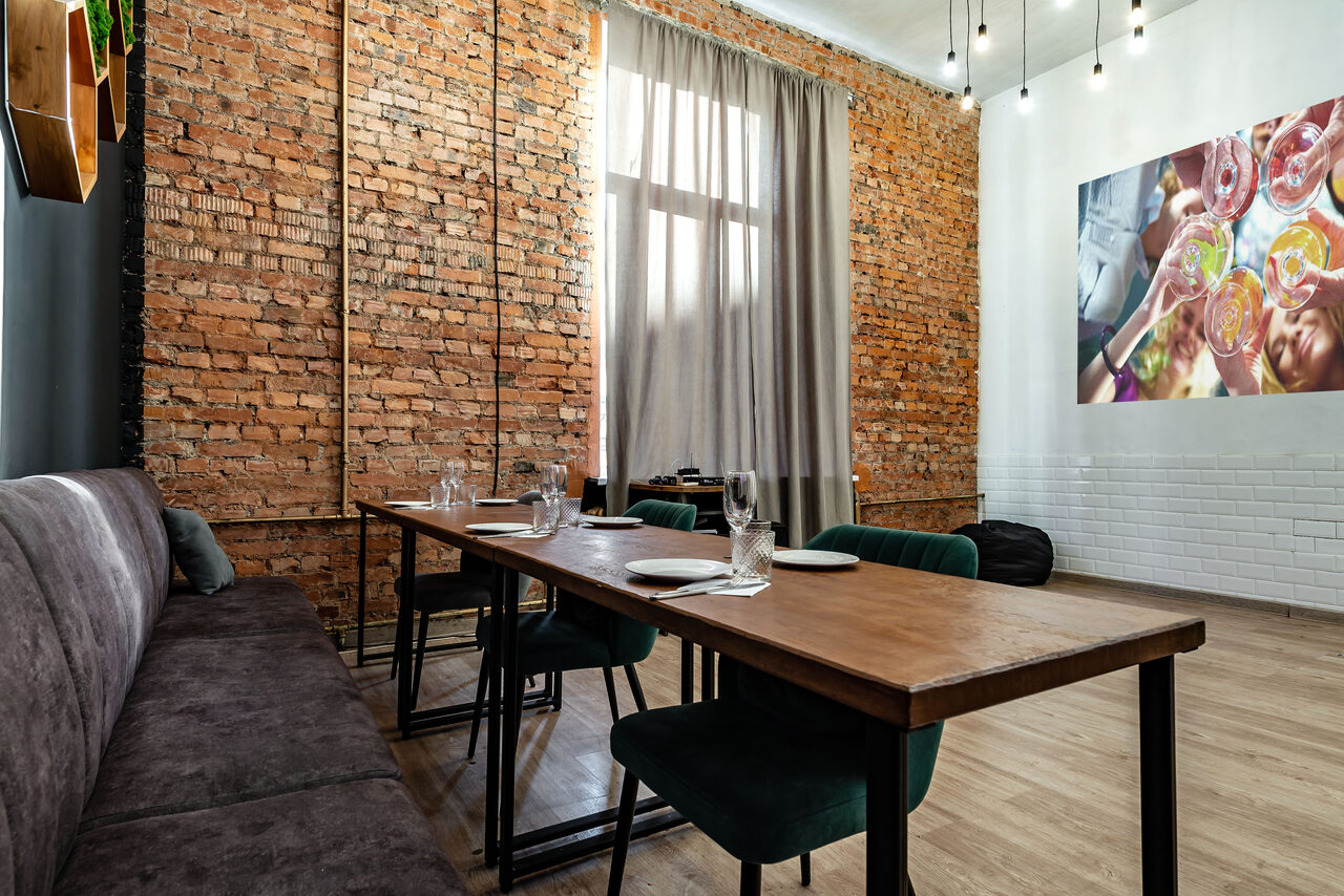 Арендовать Rubin Loft (Рубин Лофт). Лофт средний. Лофт-пространство с кирпичными стенами и деревянным полом. Длинный стол с тарелками и бокалами, по обеим сторонам - стулья и диван. Большое окно со шторами. На стене фото бокалов. Потолочные светильники на проводах.
