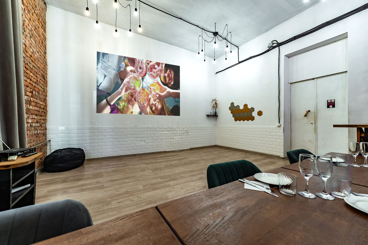 Арендовать Rubin Loft (Рубин Лофт). Лофт средний. Пространство с белыми стенами, одна из которых облицована белым кафелем и кирпичом. На стене проектируется изображение. Натяжные лампы на потолке. Стол с приборами и бокалами. Черный пуф у стены. Светлое деревянное покрытие пола.