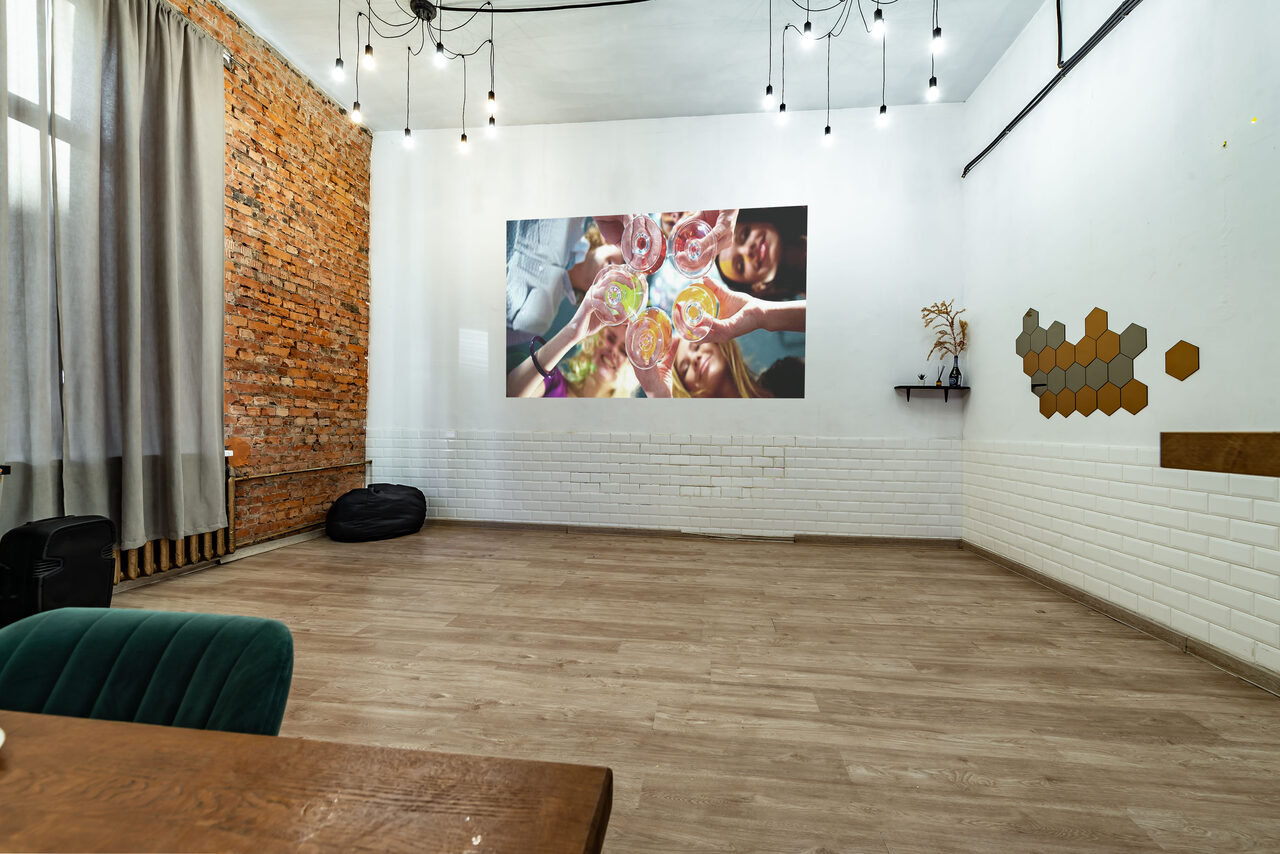 Арендовать Rubin Loft (Рубин Лофт). Лофт средний. Многофункциональное помещение с кирпичной и белой кирпичной стенами. Пол деревянный, потолок с подвесными лампами. На стене проекция изображения, рядом полки с декором. Большие серые шторы, мягкое кресло и деревянный стол.