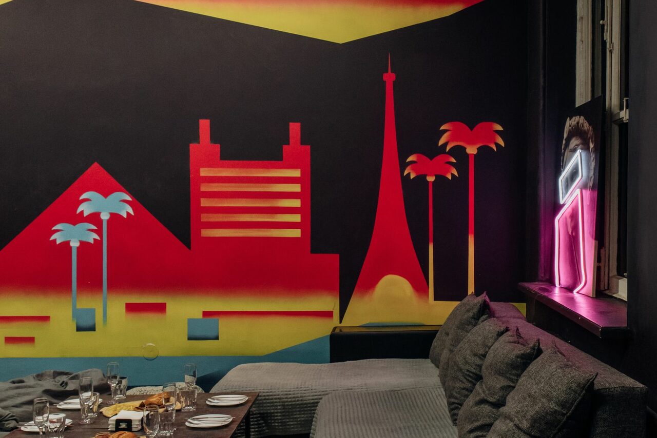 Арендовать Зал Лас-вегас. Лофт-зал в американском стиле . Комната с черными стенами, украшенными ярким граффити в виде зданий и пальм. Угловой диван с серыми подушками. Небольшой стол с посудой и напитками. Неоновая вывеска человечка рядом с окном.