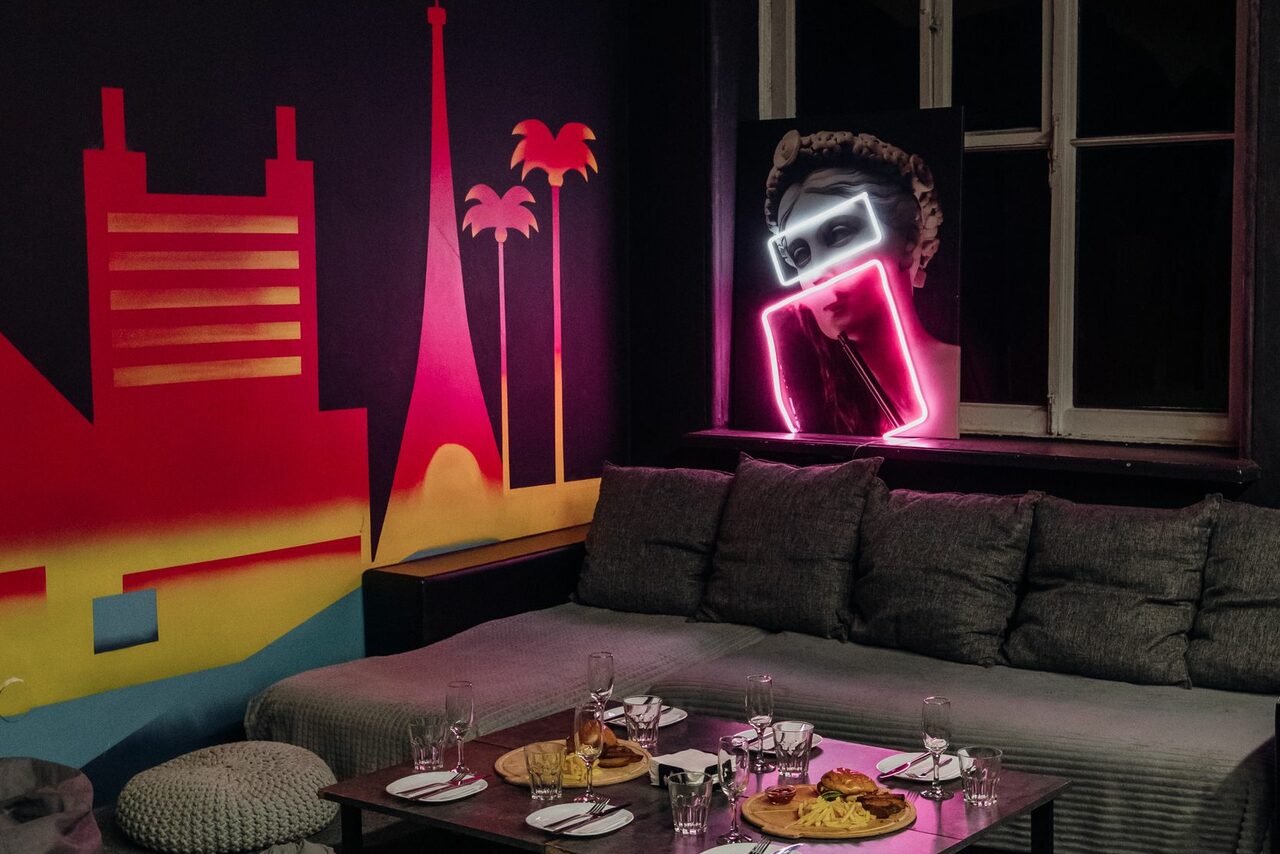 Арендовать Зал Лас-вегас. Лофт-зал в американском стиле . Лофт-пространство с мягким серым диваном и подушками. Деревянный стол с закусками и бокалами. На стене настенное граффити с абстрактными зданиями и пальмами в красно-желтых тонах. Неоновая вывеска рядом с окном. Полумрак.