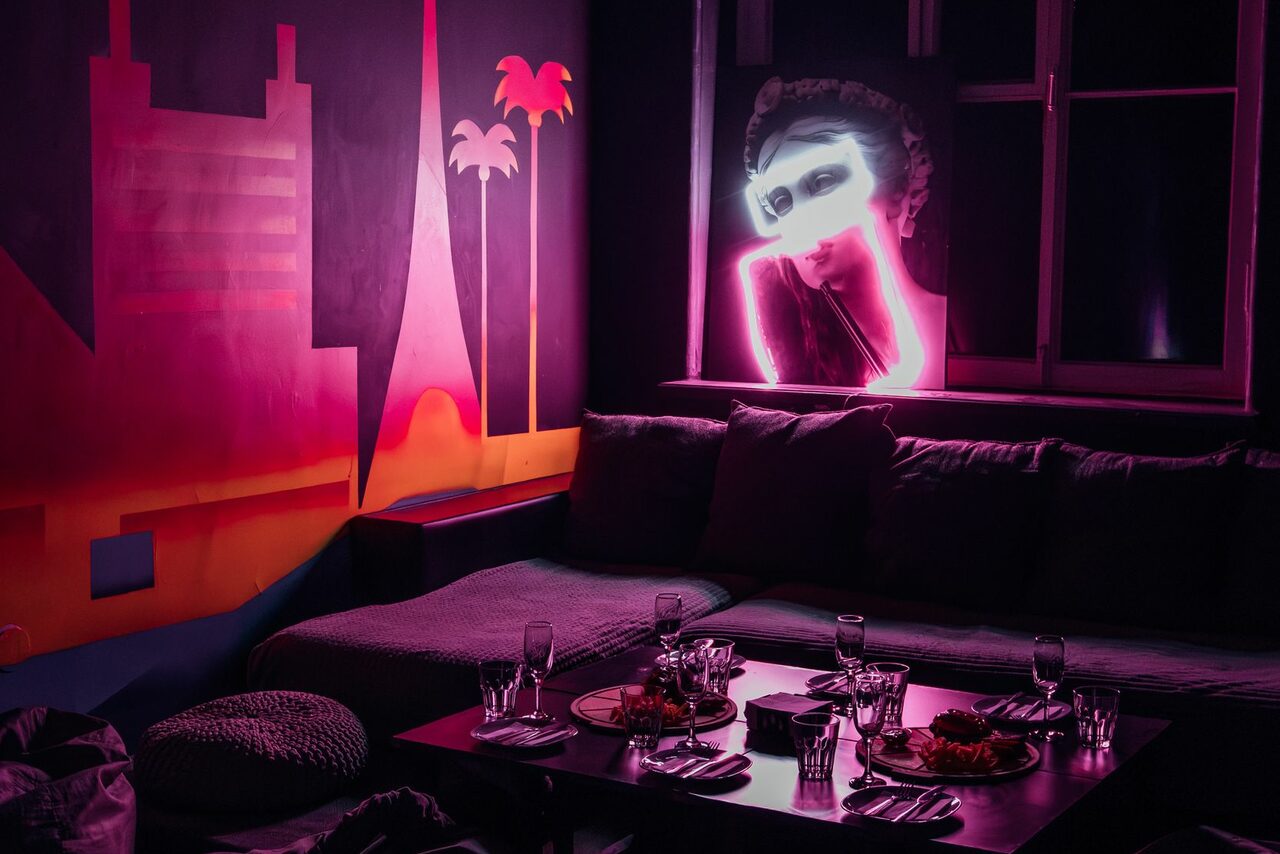Арендовать Зал Лас-вегас. Лофт-зал в американском стиле . Камерное пространство с низким столом, окружённым диваном с подушками и несколькими пуфами. На стене яркий неоновый арт с изображением лица. Расставлены бокалы и тарелки. Освещение приглушённое с розовым неоновым акцентом.