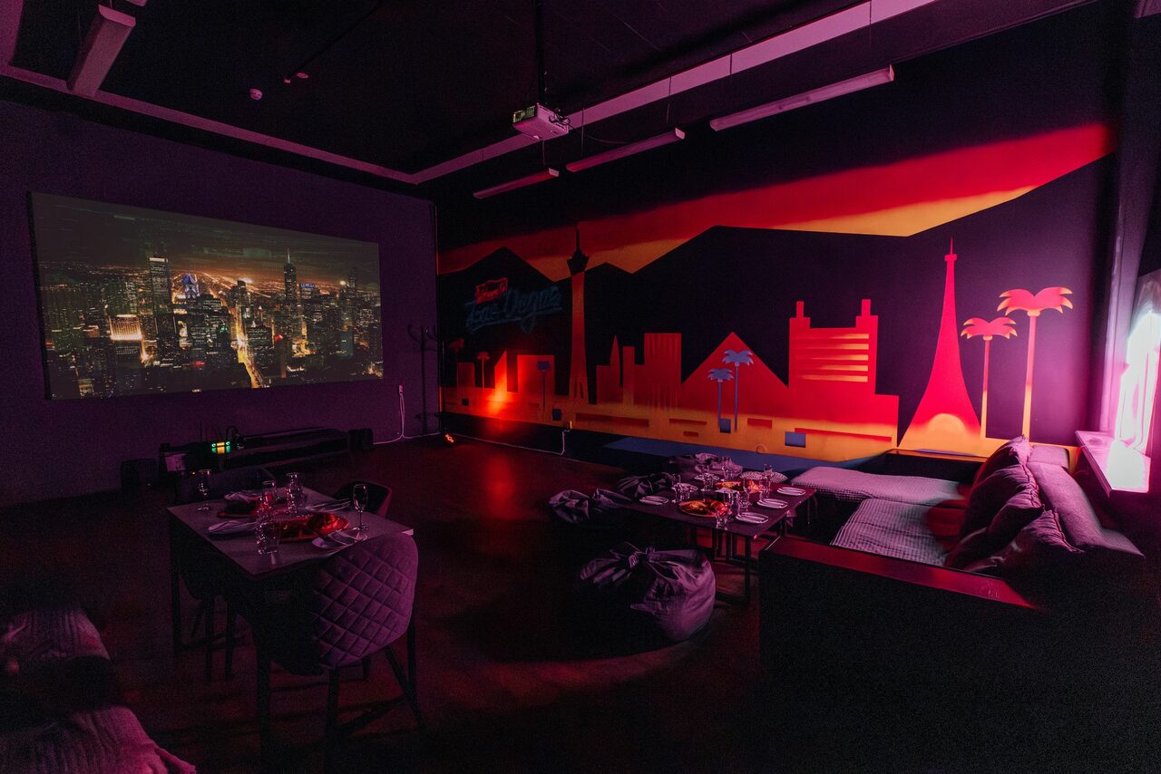 Арендовать Rubin Loft (Рубин Лофт). Зал Лас-вегас. Темная комната с проектором, показывающим изображение мегаполиса. На стене красочный настенный рисунок в виде городского пейзажа. Мебель: столы и стулья, диван, подушки. Тусклое освещение, теплые тона.