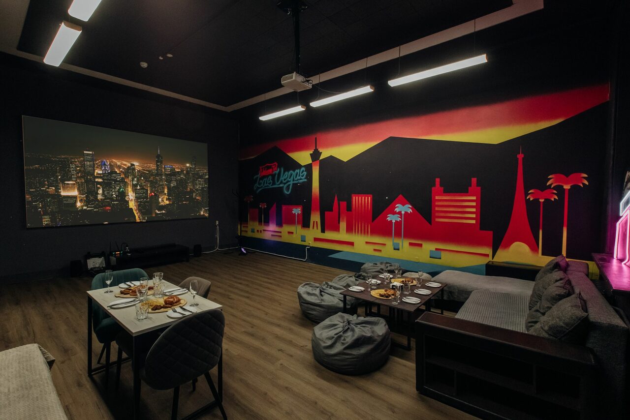 Арендовать Rubin Loft (Рубин Лофт). Зал Лас-вегас. Комната с темными стенами и деревянным полом. На стене яркий городской пейзаж. Большой экран с изображением города. Обеденный стол со стульями, диван, мягкие кресла и обилие подушек. Подвесные светильники.