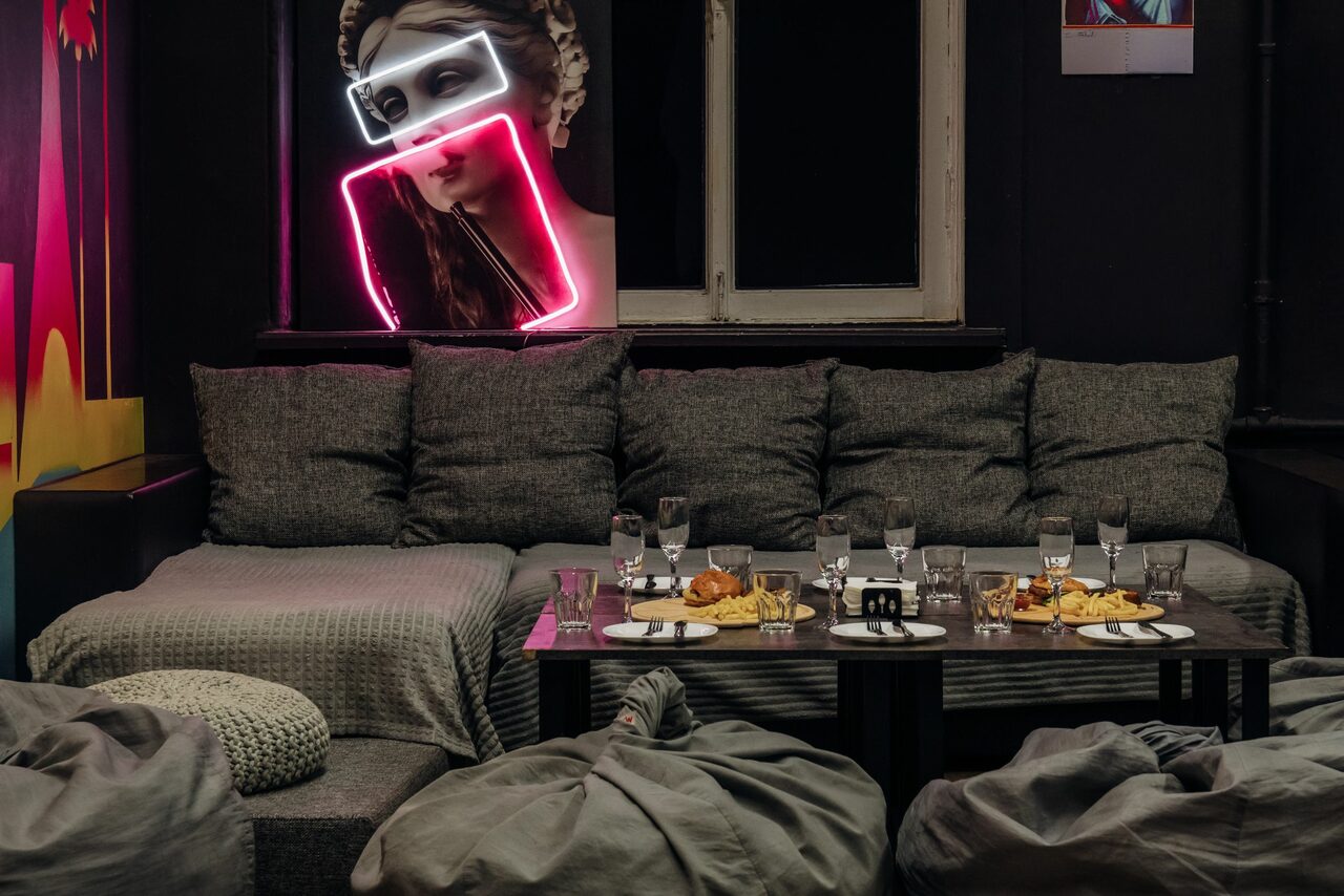 Арендовать Rubin Loft (Рубин Лофт). Зал Лас-вегас. Современное пространство с серыми диванами и пуфами, низким столом с закусками и напитками. На стене неоновая женская фигура и яркий арт. Тёмные стены, окно с белой рамой. Интимное освещение.