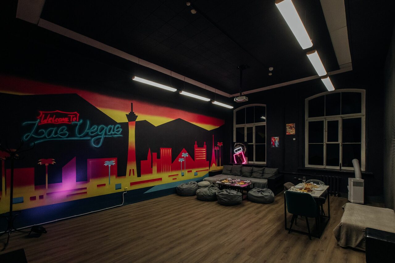Арендовать Rubin Loft (Рубин Лофт). Зал Лас-вегас. Арт-пространство с неоновым граффити на стене. Небольшие мягкие кресла и столы с закусками. Диван, стулья, длинные потолочные светильники. Темный потолок, деревянный пол, большие окна. Настенное искусство с городским пейзажем и неоновые вывески.