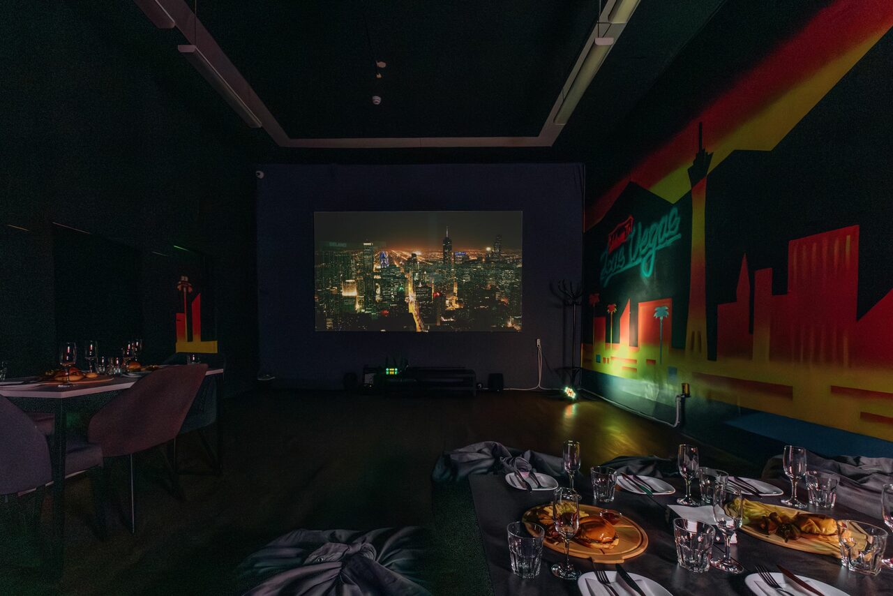Арендовать Rubin Loft (Рубин Лофт). Зал Лас-вегас. Просторная затемненная комната с проектором, показывающим городской пейзаж. На стене граффити с силуэтами зданий. Расставлены столы, сервированные блюдами и стеклянной посудой. Стены темные, пол деревянный, освещение приглушенное.