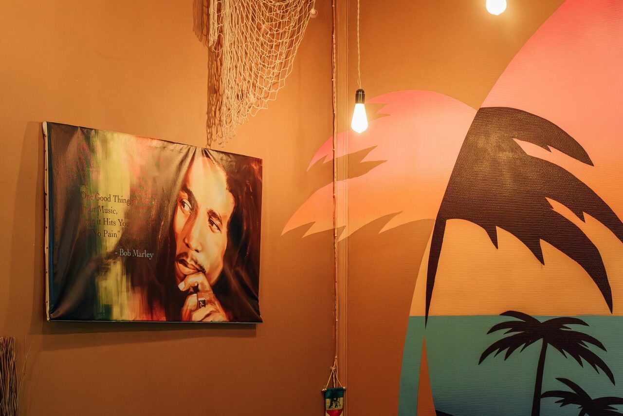 Арендовать Зал Ямайка. Лофт с большим диваном и экраном. Стена с настенными искусствами: плакат с изображением человека и цитатой, декоративная сетка, яркое фреска с пальмами и закатом. Освещение в виде подвесных ламп, теплые коричневые и оранжевые тона.