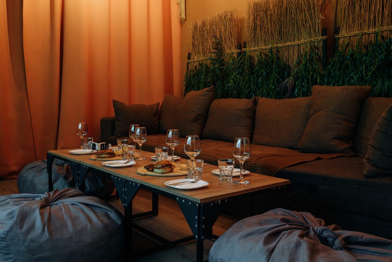 Арендовать Зал Ямайка. Лофт с большим диваном и экраном. Комната с мягким диваном и пуфами, деревянный стол с посудой и стаканами, на стене висят декоративные растения, пол деревянный, освещение тёплое, шторы оранжевые.