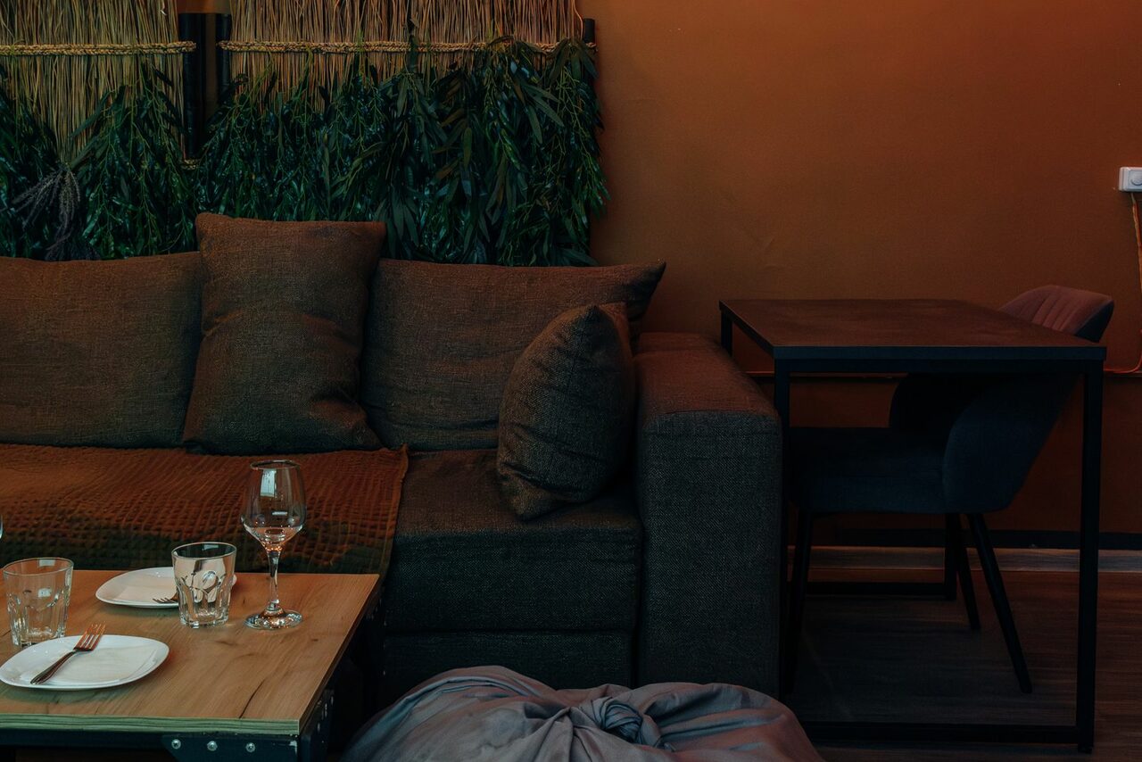 Арендовать Зал Ямайка. Лофт с большим диваном и экраном. Пространство с коричневым диваном и квадратным столом на двоих, черные стулья. Декоративная стена с зеленью. На деревянном столике бокал для вина, стаканы, тарелки с приборами. Теплое освещение, оранжево-коричневые стены, мягкие текстуры.