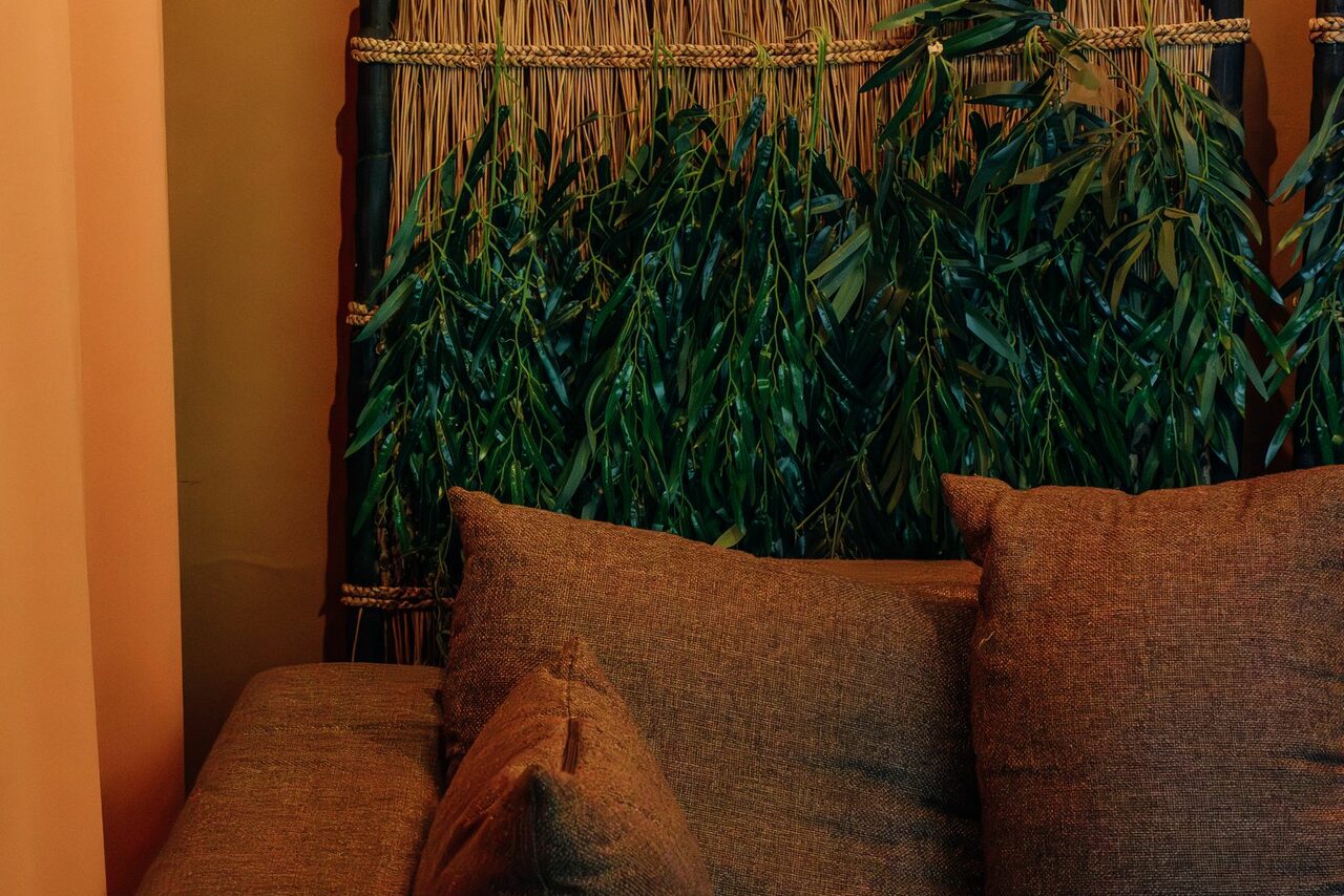 Арендовать Зал Ямайка. Лофт с большим диваном и экраном. Диван с коричневыми подушками перед декоративной панелью с зелеными листьями и плетением. Стены теплых бежевых тонов. Мягкое освещение создает уютную атмосферу.