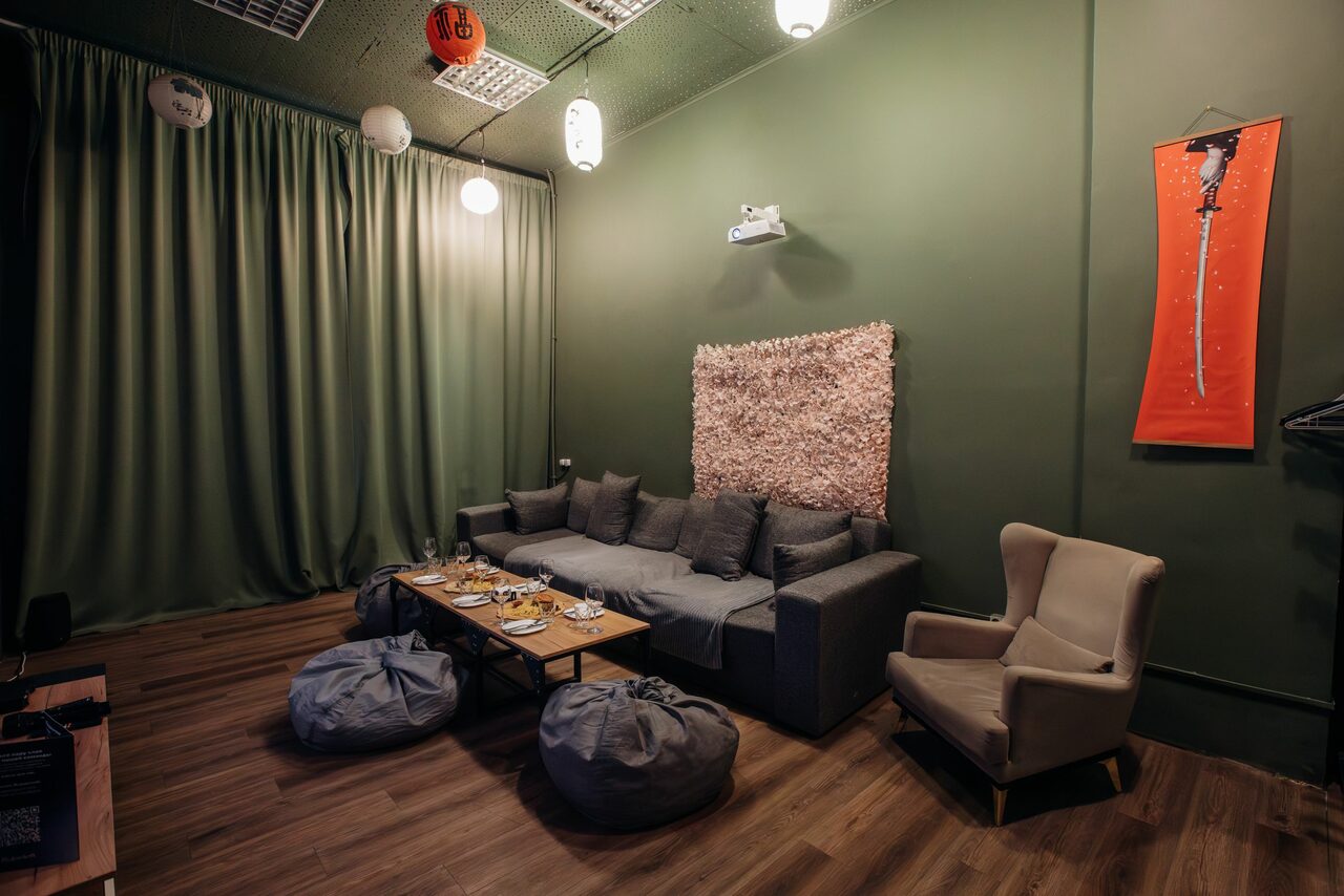 Арендовать Зал Китай. Лофт-зал в китайском стиле . Зал с зелеными стенами и занавесками, деревянный пол, серый диван и кресло, подушки-мешки, деревянный стол с посудой, настенный проектор, декоративная панель. Потолочные светильники, подвесные бумажные лампы. Красный вертикальный постер на стене.