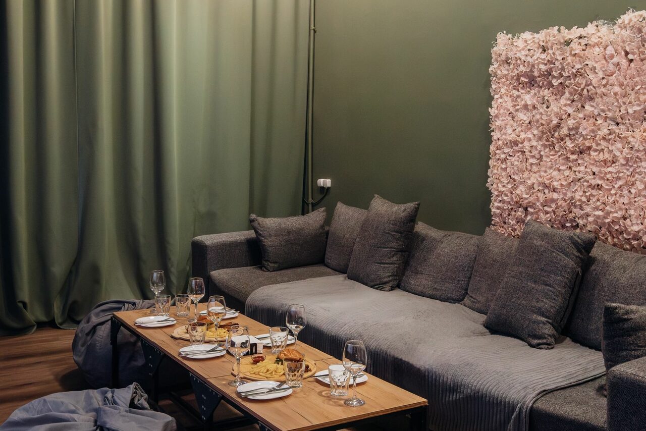 Арендовать Зал Китай. Лофт-зал в китайском стиле . Гостиная зона с серым диваном, перед ним деревянный стол с накрытыми сервизами и бокалами. Справа стена с розовыми цветочными элементами. Зелёные занавески и деревянный пол. Мягкое освещение.