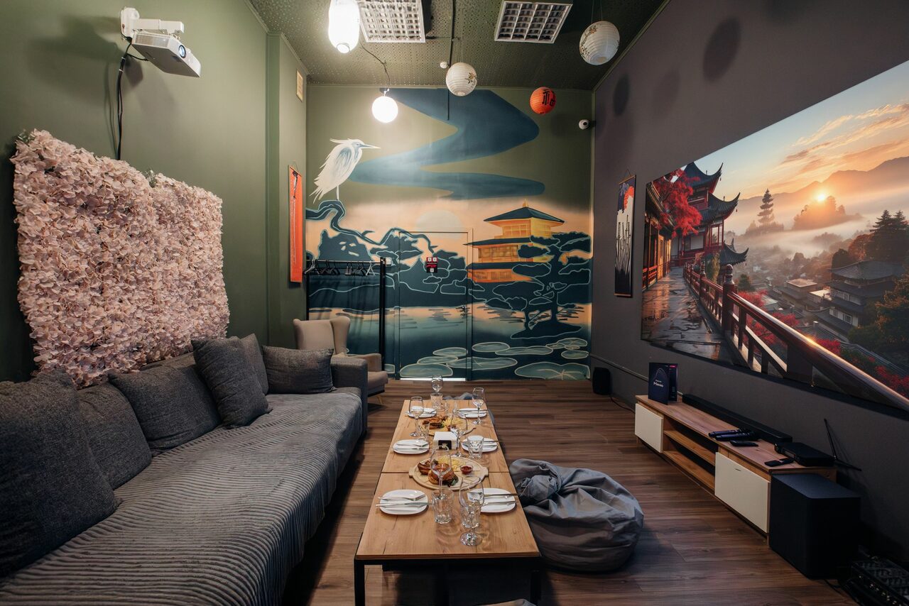 Арендовать Зал Китай. Лофт-зал в китайском стиле . Многофункциональное пространство с длинным серым диваном, журнальным столиком с едой и напитками, креслом, пуфом. На стене большая роспись с японским пейзажем. Проектор на потолке, полосатый деревянный пол, подвесные светильники, стены зелёного и серого цвета.